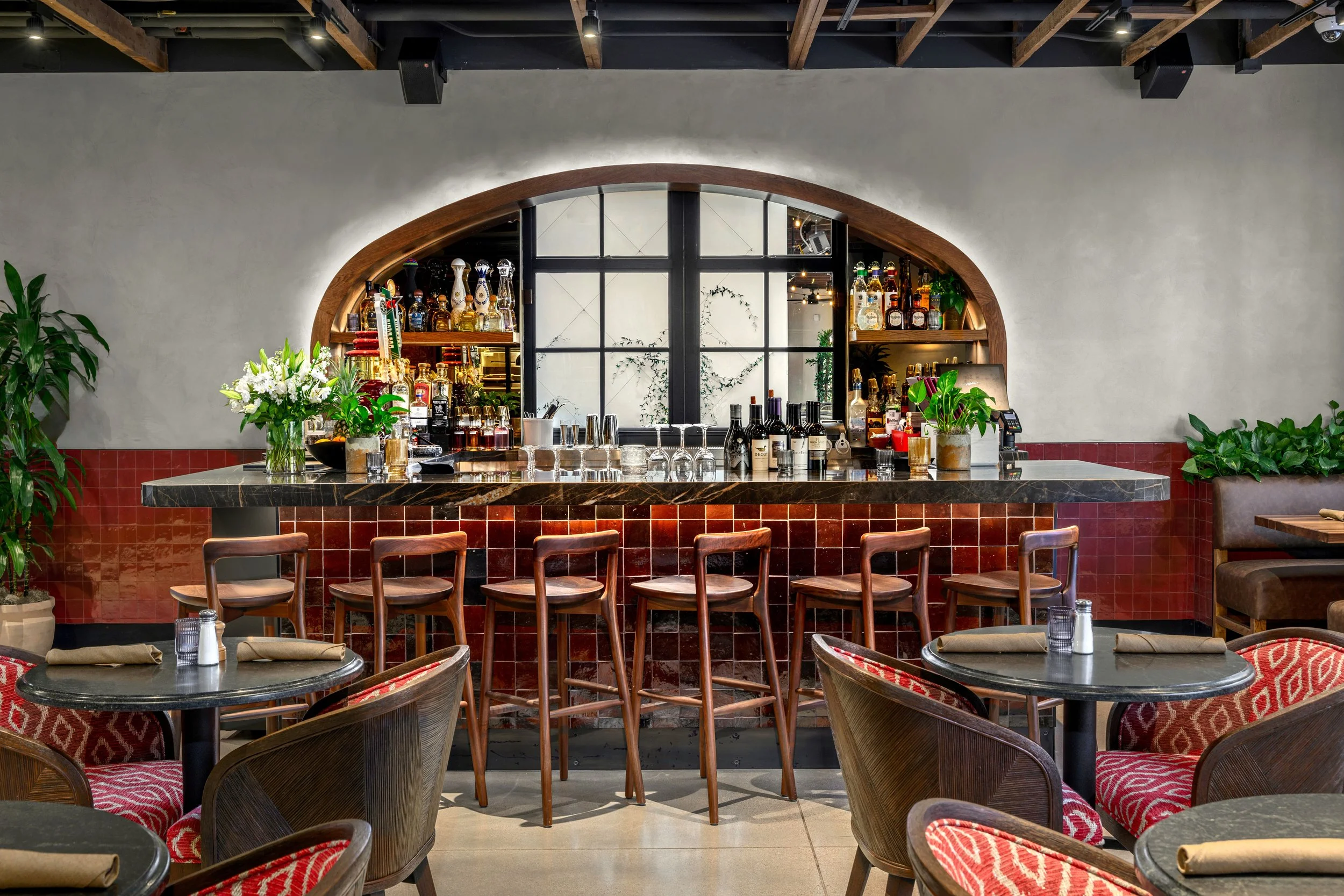 Interior of a restaurant bar area with a curved bar counter, wooden high chairs, tables with chairs, and decorative plants. The bar has bottles of alcohol and glasses, with a window behind it.
