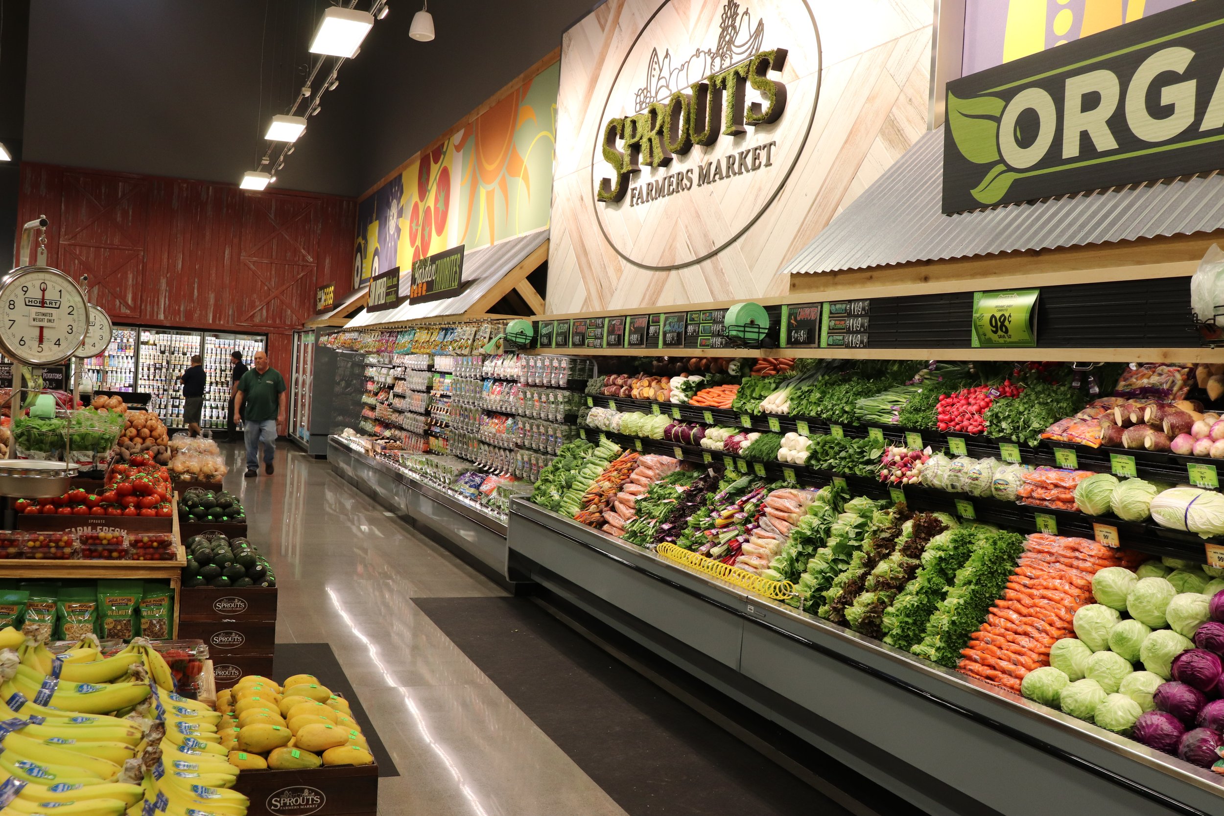Fresh vegetables and fruits on display in a farmer's market with a sign reading 'Sprouts Farmers Market' on the wall.