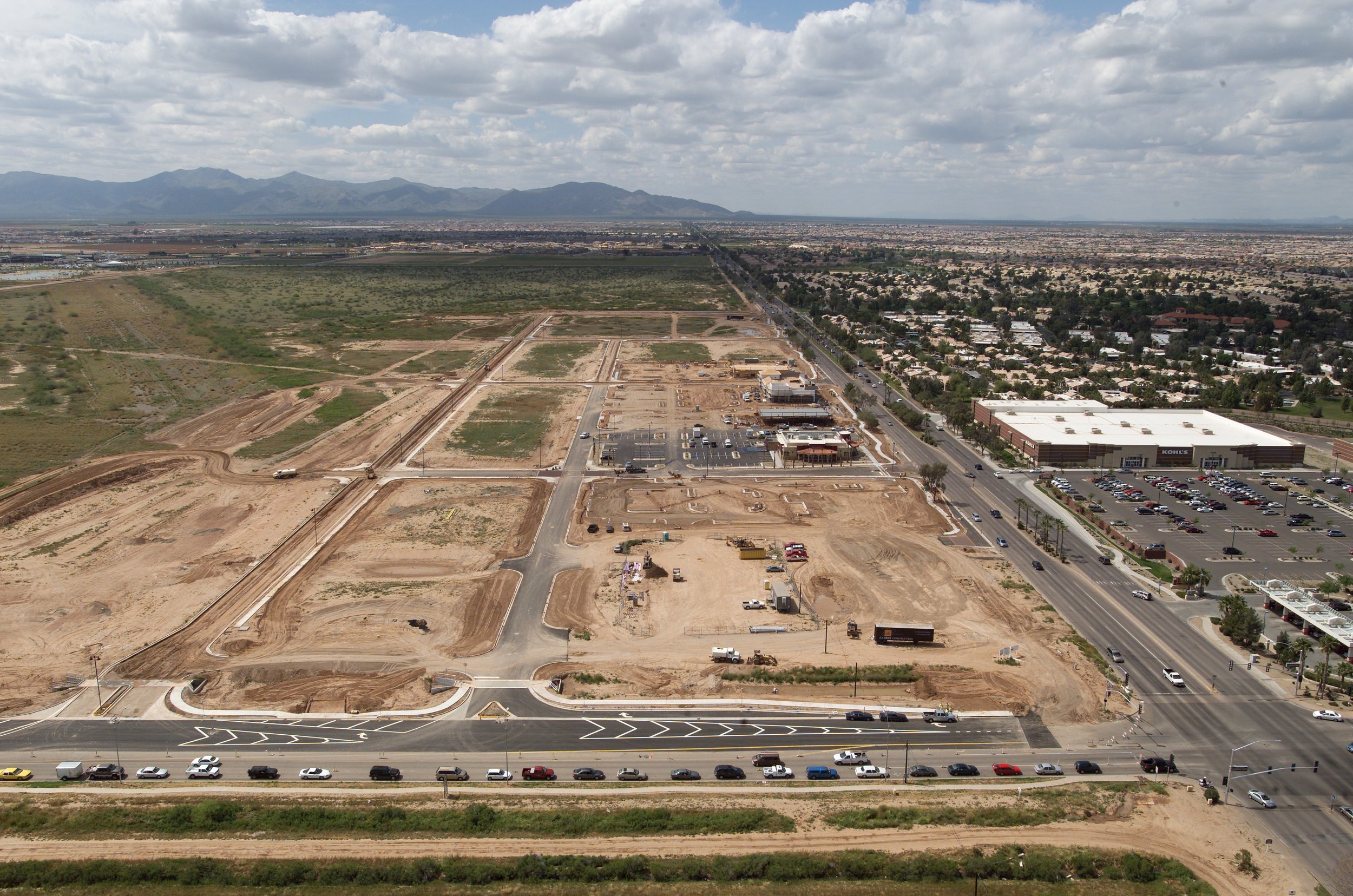 Aerial view of a construction site with dirt roads, partially built structures, and parked vehicles, next to developed commercial area and a busy street with moving cars in an urban area with mountains in the background.