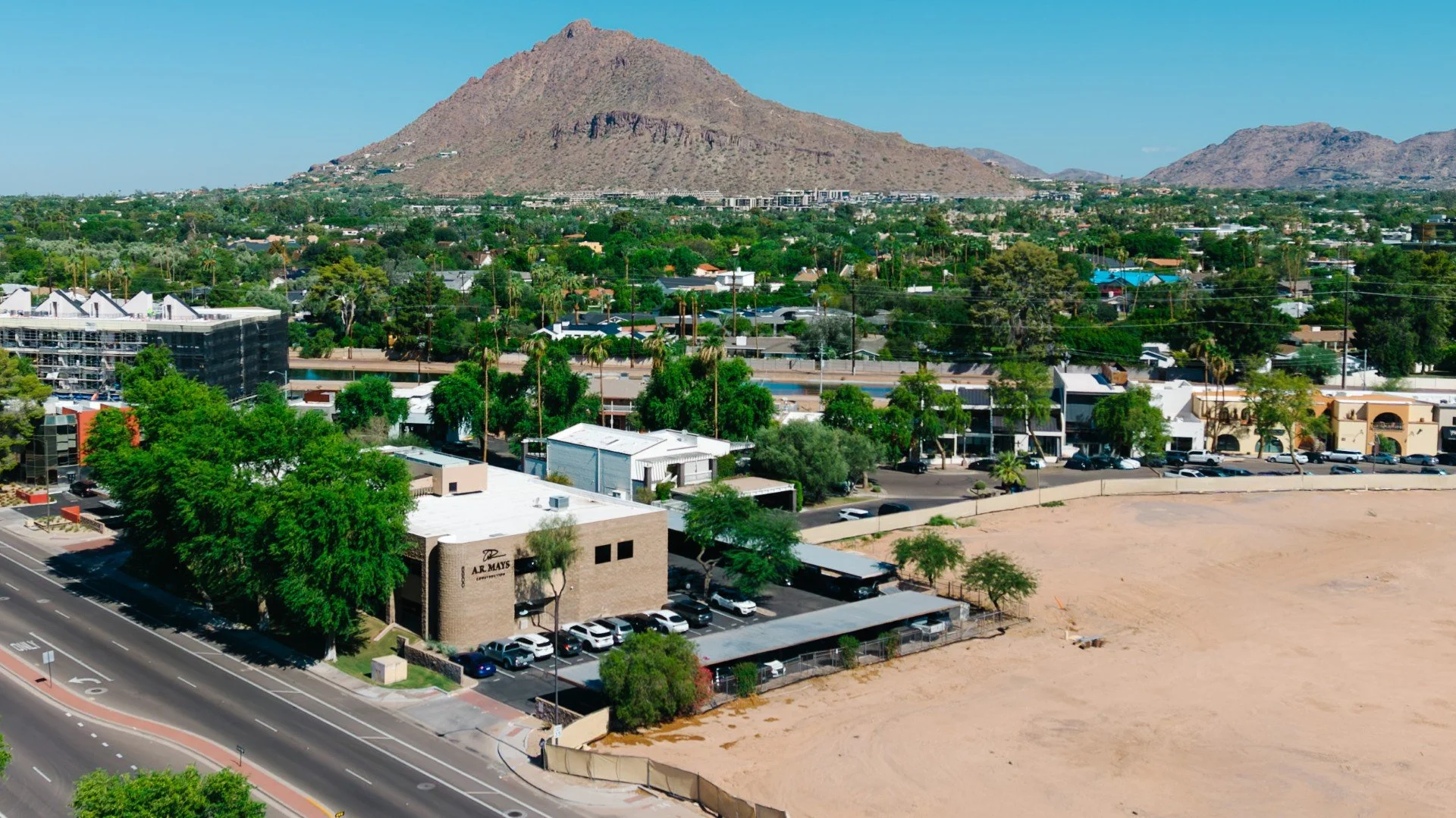 Aerial view of a cityscape with apartment buildings, commercial structures, and a parking lot, with a dry, sandy area in the foreground, and a large mountain in the background under a clear blue sky.