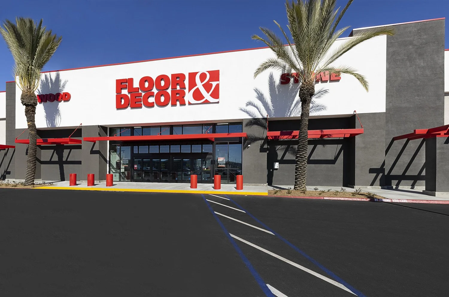 Exterior view of a flooring and decor store with a large white sign and red lettering, palm trees, and a parking lot with painted parking lines and red parking bumpers.