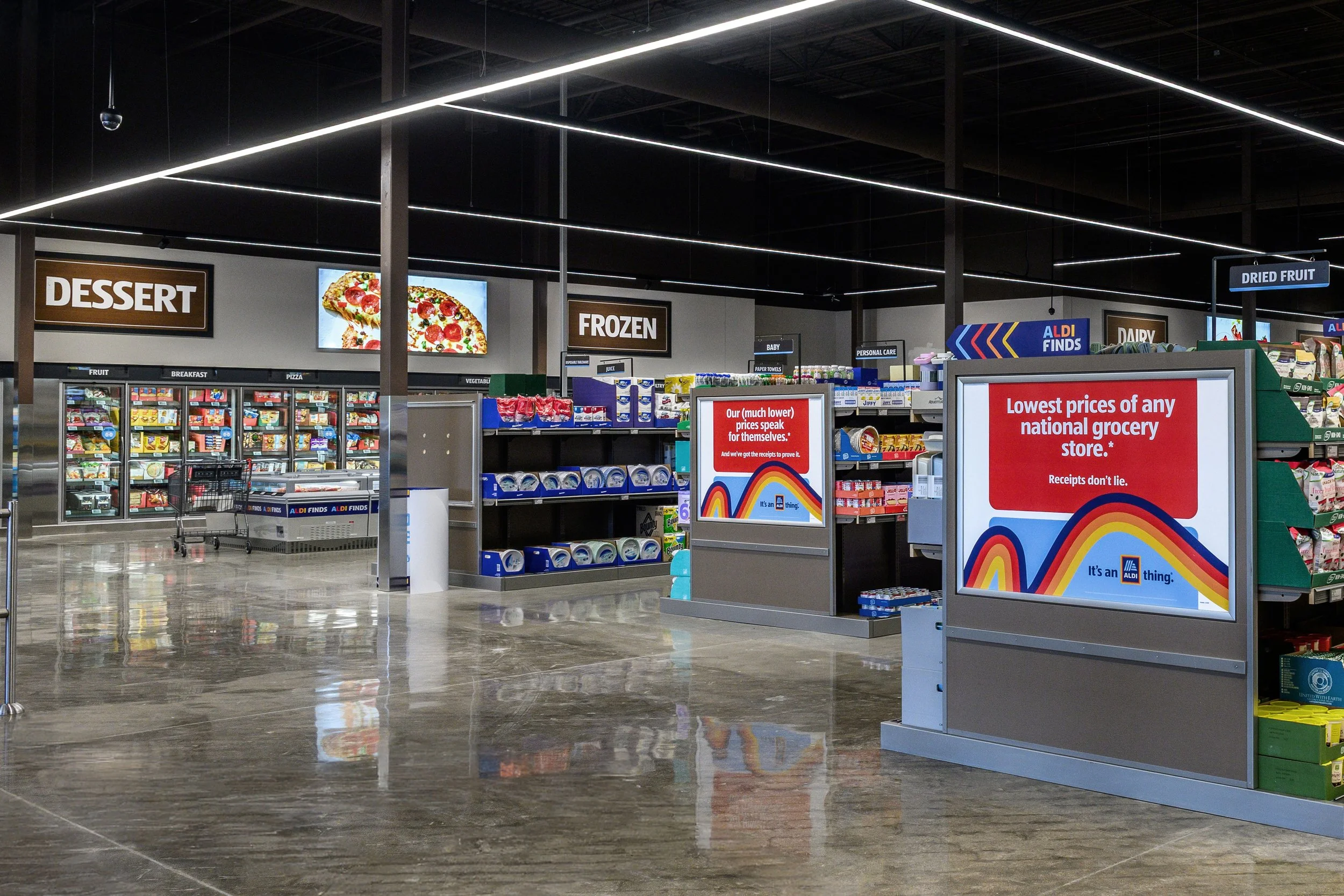 Interior of an Aldi grocery store, showing dairy and frozen food sections, signs for dessert, frozen, and dried fruit, and display screens with promotional messages.