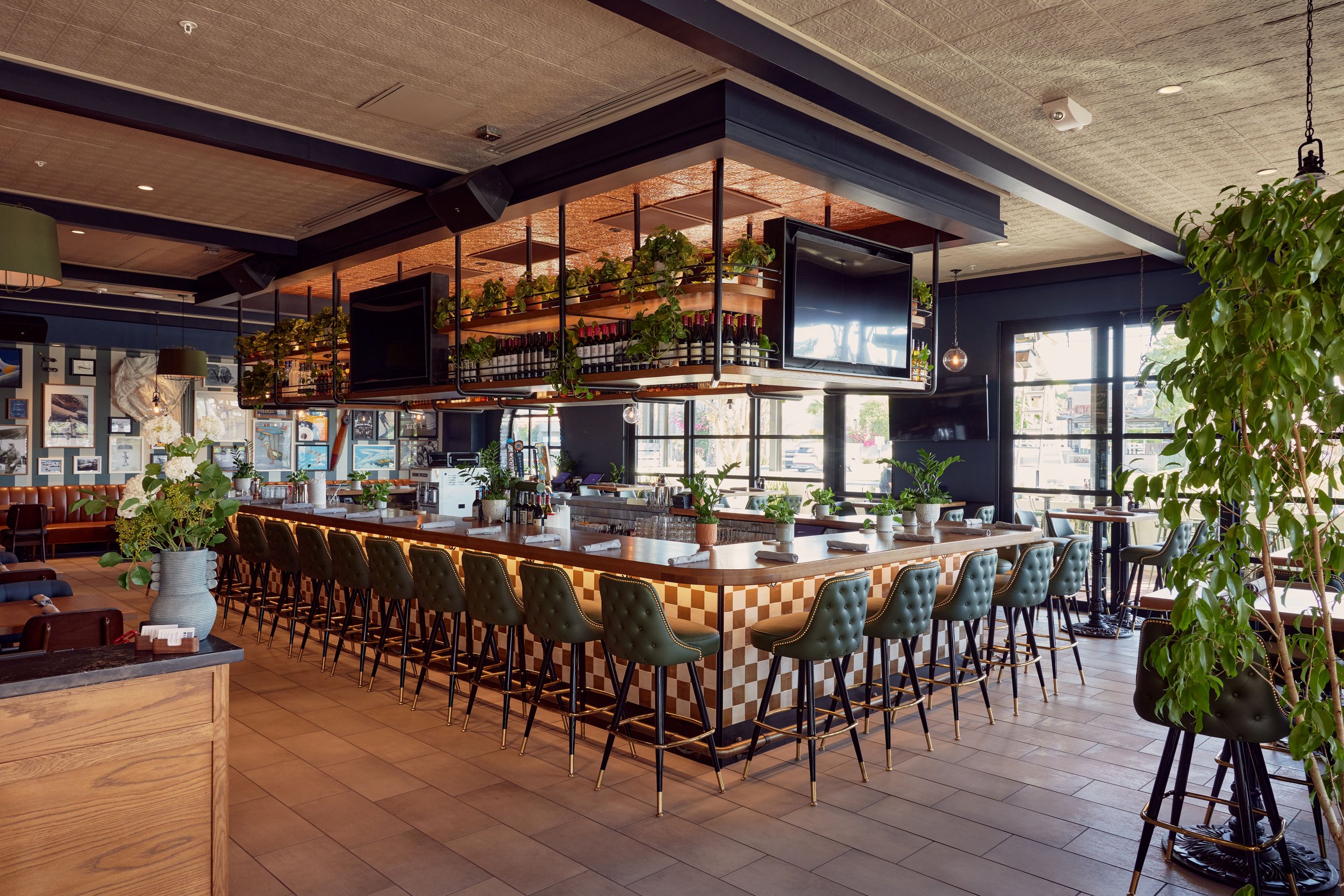 Inside a modern restaurant with a central bar, green upholstered barstools, potted plants, and a wall decorated with framed pictures.