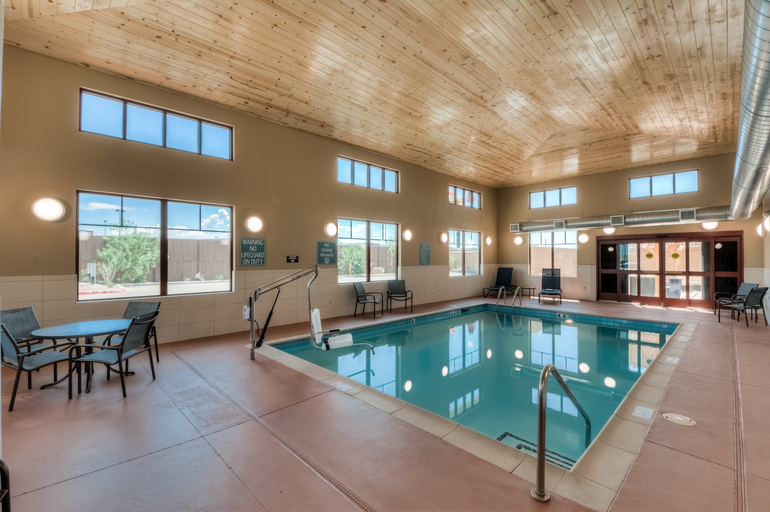 Indoor pool area with chairs, tables, and large windows, wooden ceiling, and brick doors.