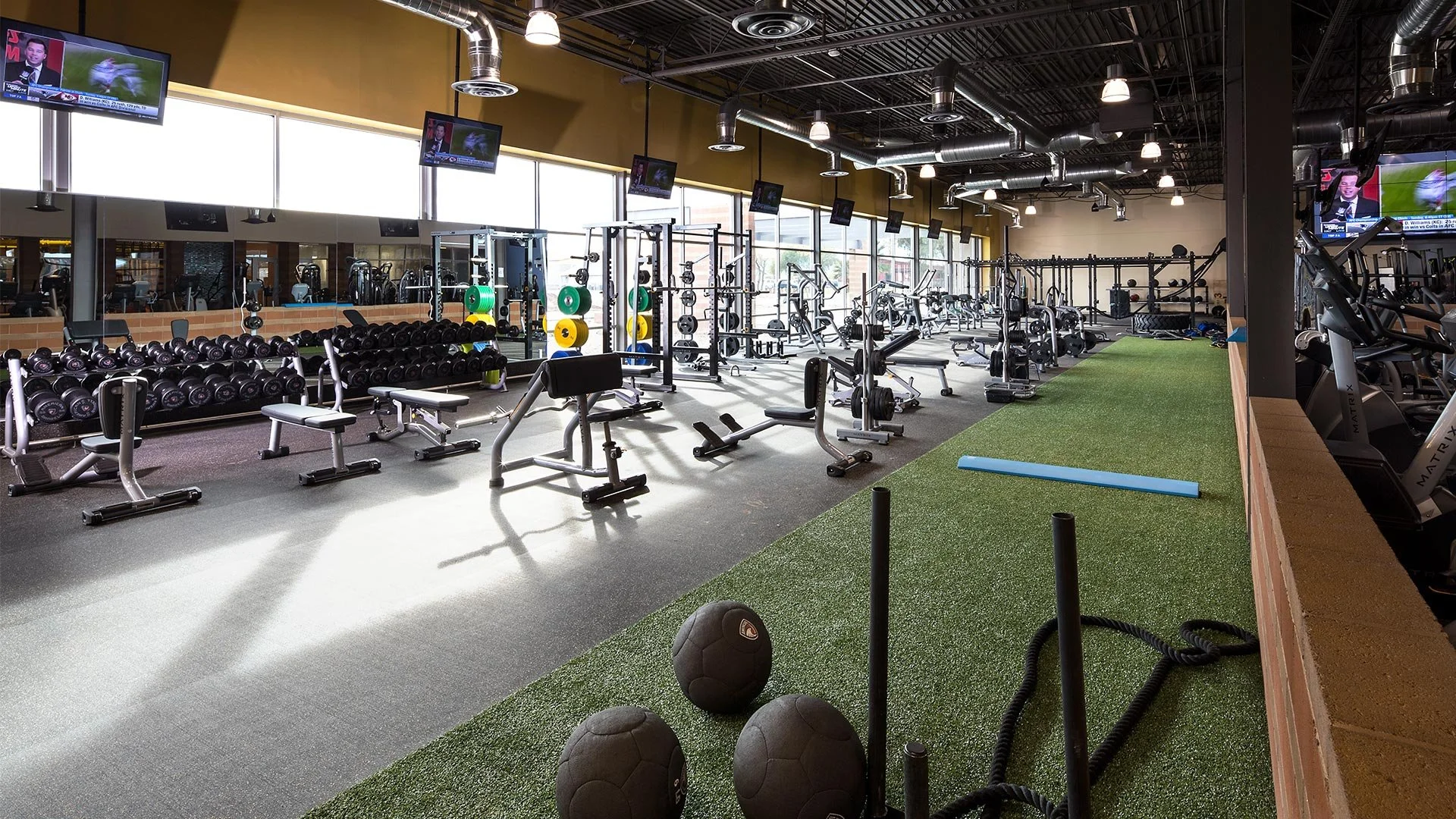 A spacious gym with various workout equipment, including dumbbells, weight machines, and cardio machines, with large windows, overhead lighting, and a green turf area with medicine balls and battle ropes.