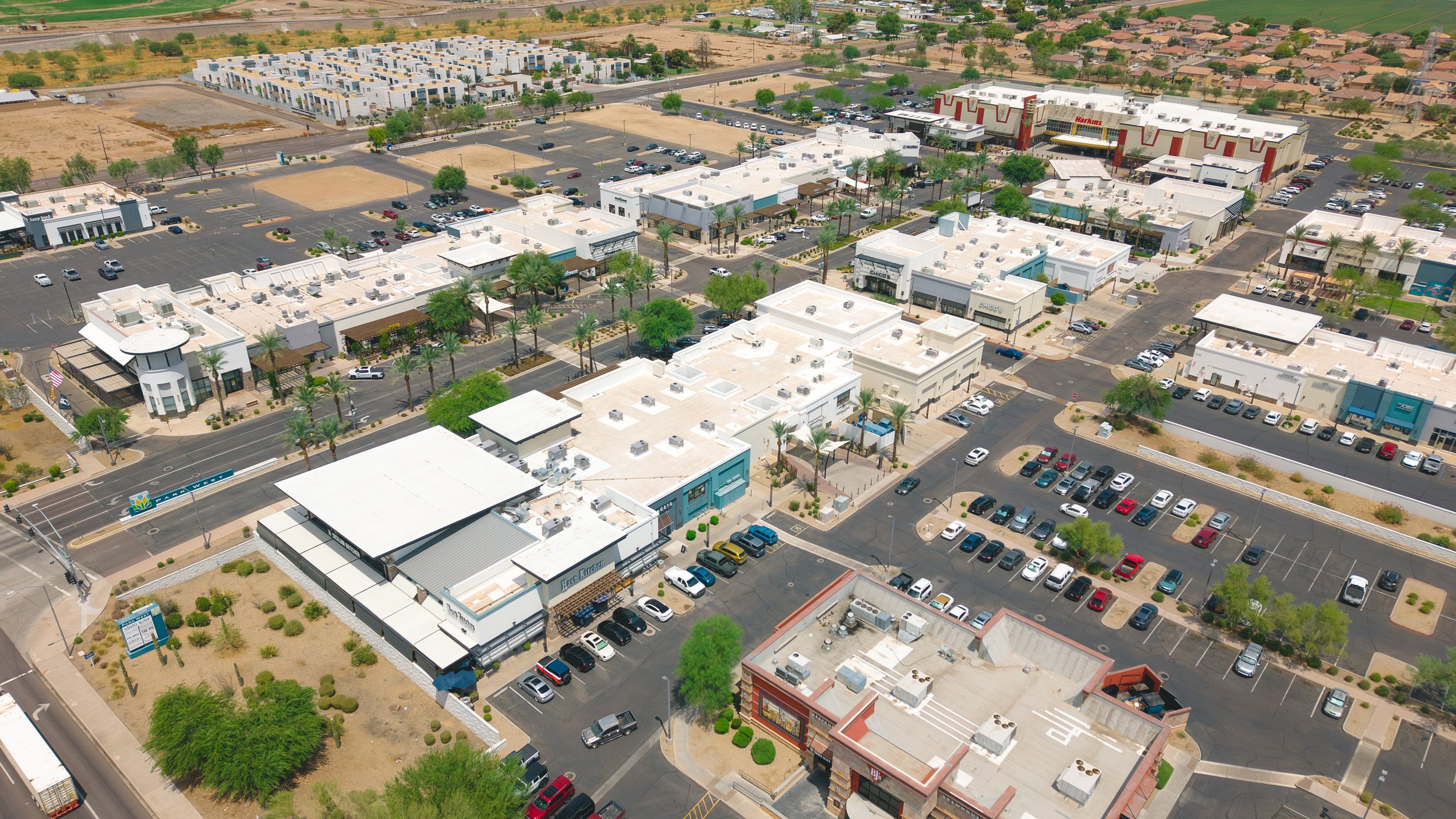 An aerial view of a shopping plaza with multiple stores, parking lots with cars, and surrounding desert landscape with sparse trees.