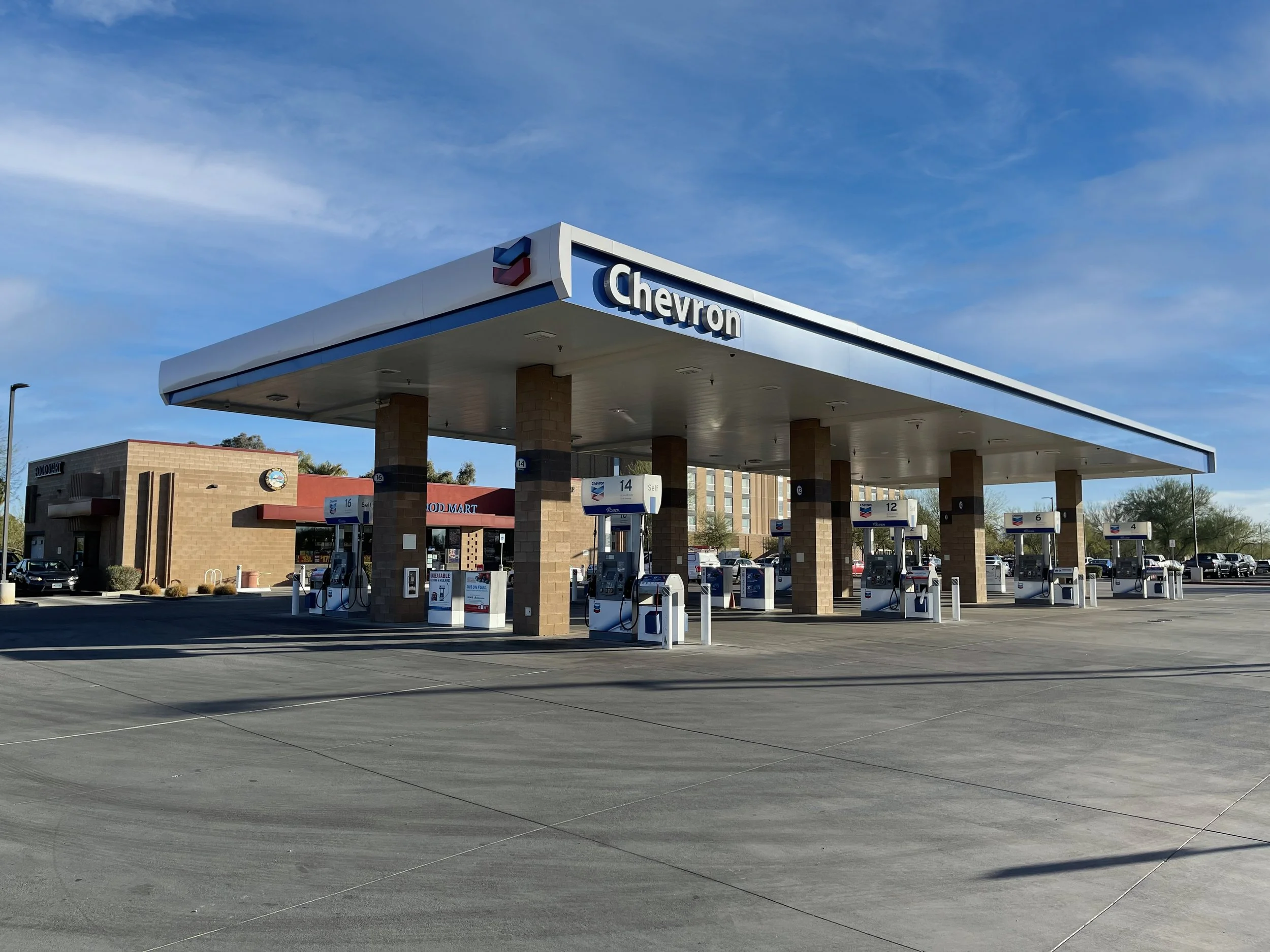A Chevron gas station with six fueling pumps under a large canopy, and a convenience store in the background on a clear day.