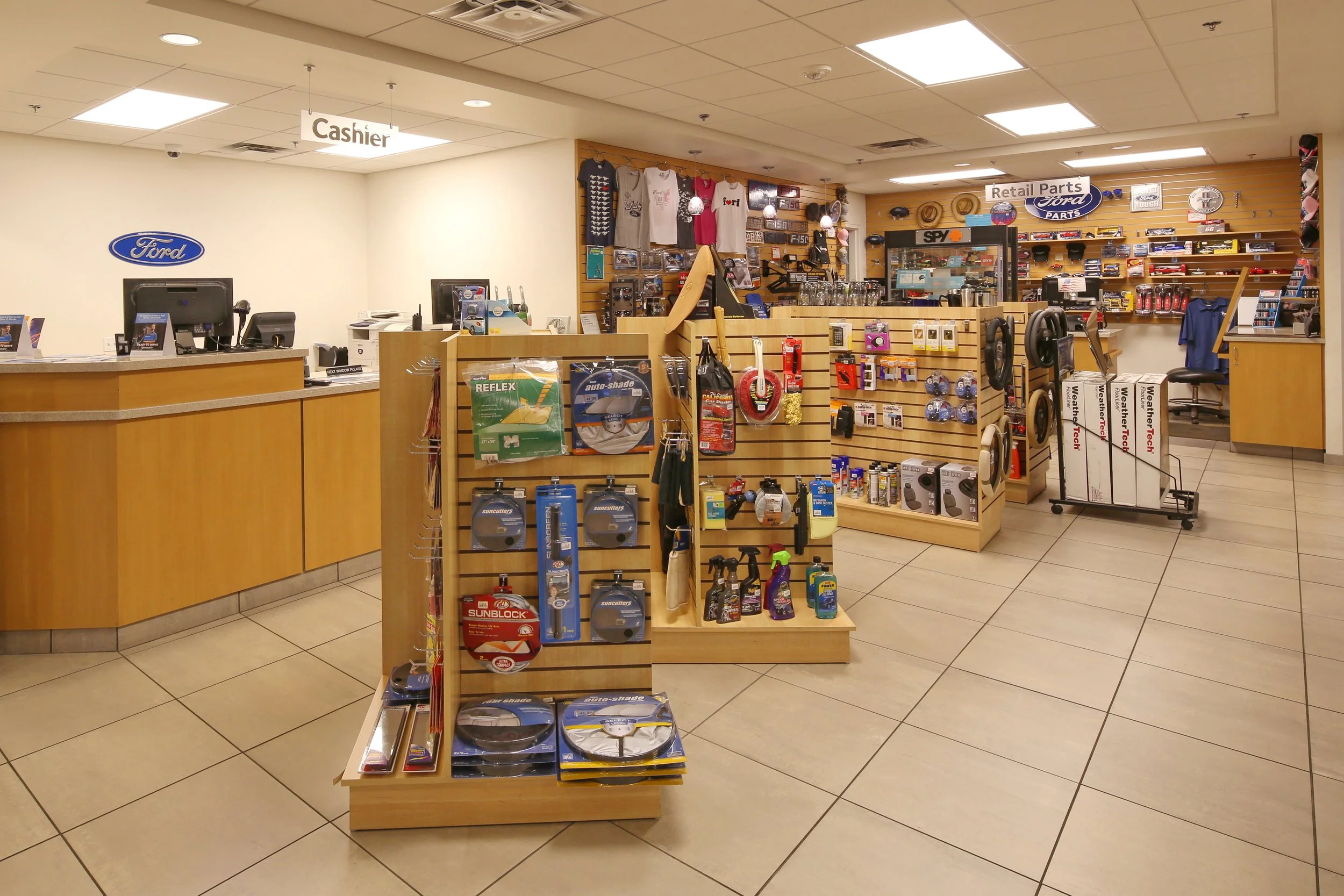 Inside a Ford dealership with the cashier desk on the left and automotive accessories and parts on display shelves in the middle and back. Signs for "Cashier" and "Retail Parts" are visible.