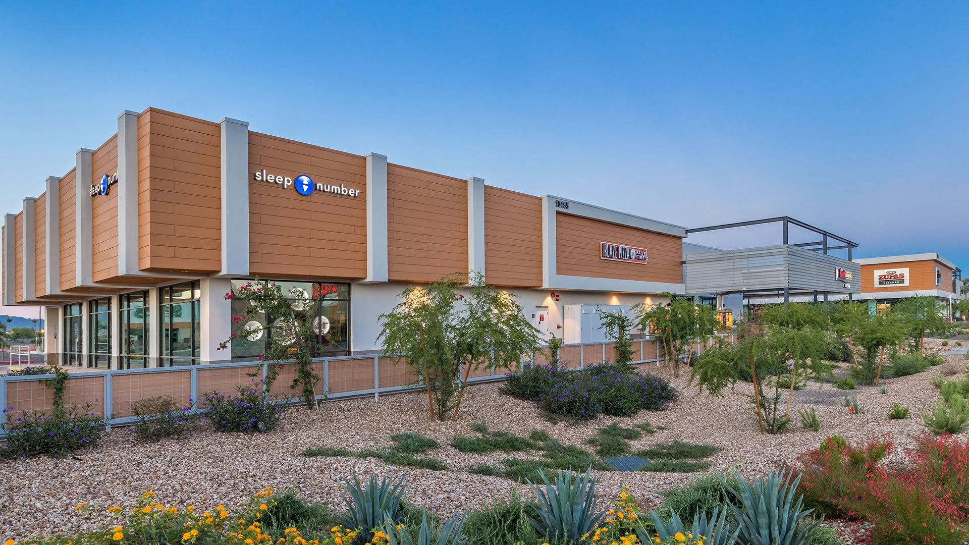 Exterior view of a shopping center with storefronts for Sleep Number, Blaze Pizza, Zuppa's, and a pretzel shop, surrounded by desert landscaping with various plants and small trees under a blue sky.
