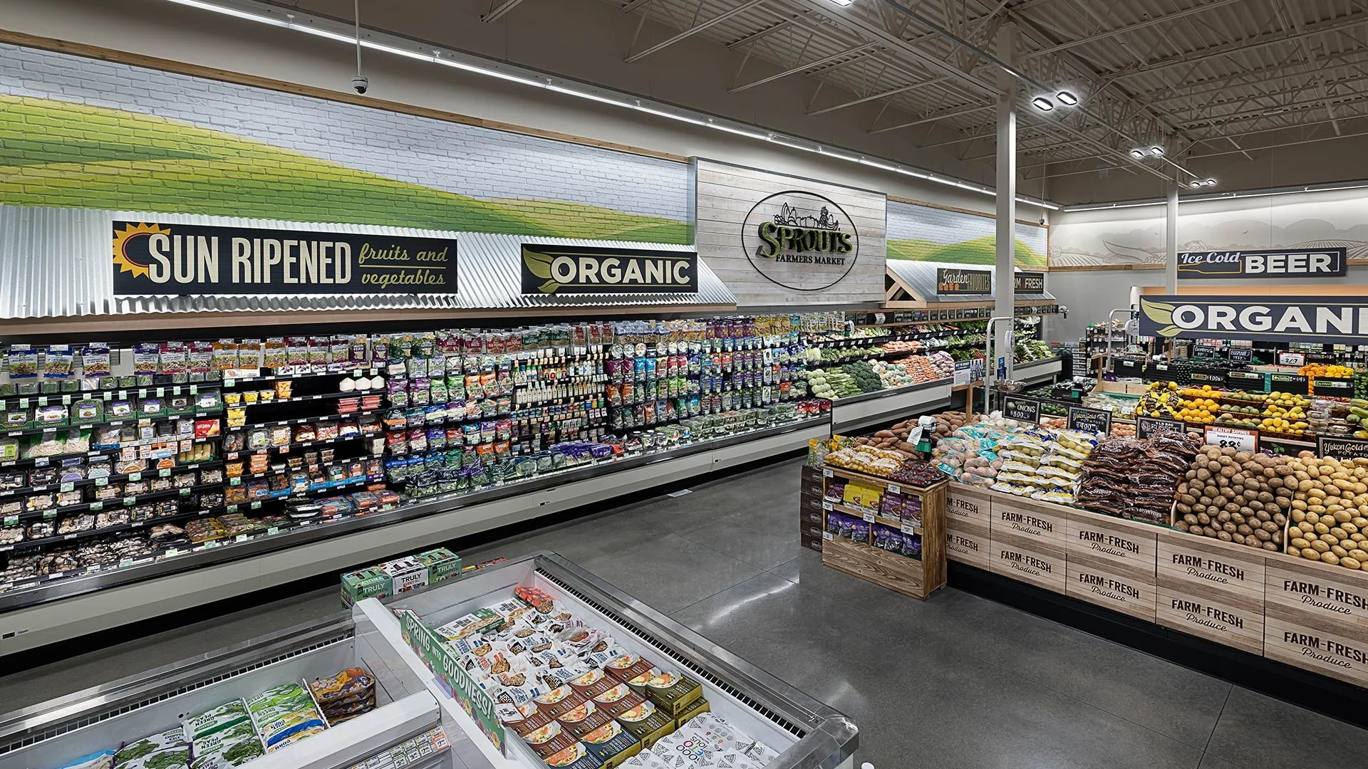 Interior of a grocery store aisle with produce section, featuring signs for Sun Ripened fruits and vegetables, organic products, and other fresh items, with shelves of packaged goods and fresh produce on display.