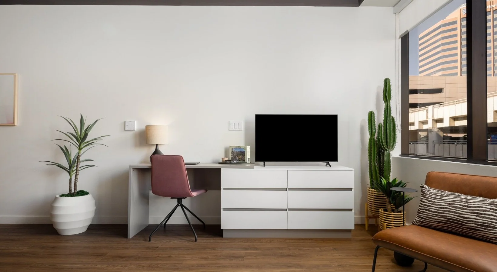 Living room with a white desk, pink chair, a lamp, a flat-screen TV, a large potted cactus, and a sofa with striped pillows near large window showing a cityscape.
