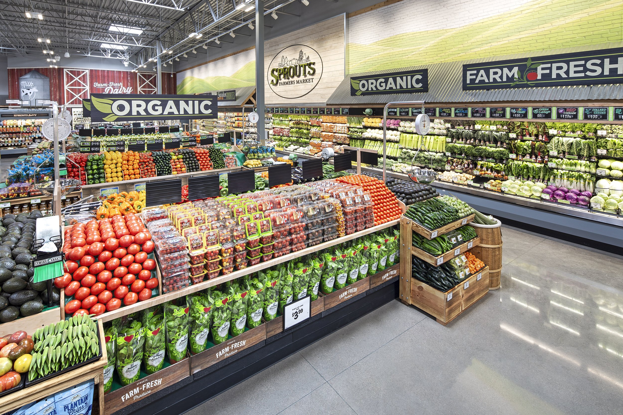 A grocery store produce section with fresh vegetables and fruits arranged in display bins and crates, signage indicating organic and farm-fresh products, and a branded sign for Sprouts Farmers Market.