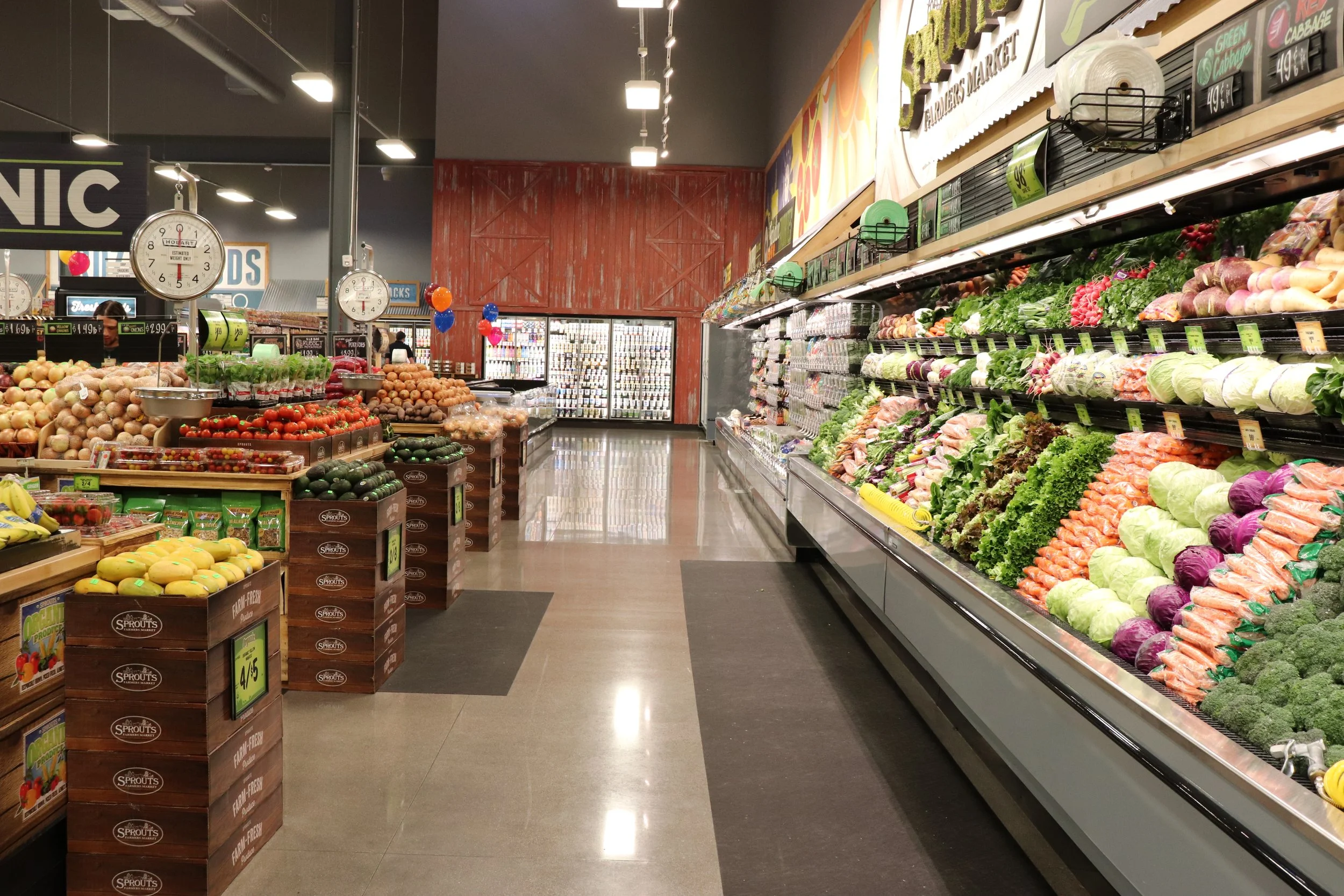 Inside a grocery store with produce section on the right displaying various vegetables like lettuce, carrots, purple cabbage, broccoli; on the left, stacks of fruits including bananas, tomatoes, and onions, with hanging clocks and balloons in the bac