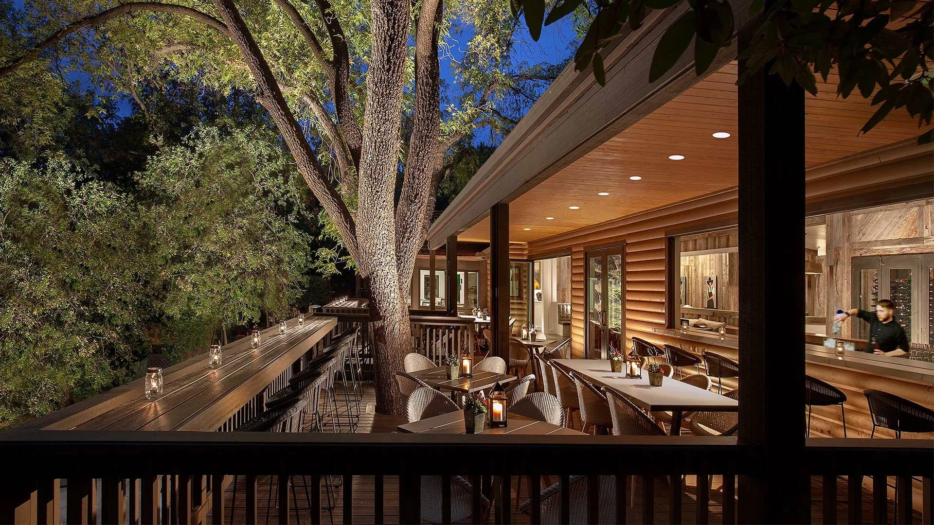 Nighttime outdoor dining area on a wooden deck with tables, chairs, candles, and a large tree, adjacent to a building with warm lighting.