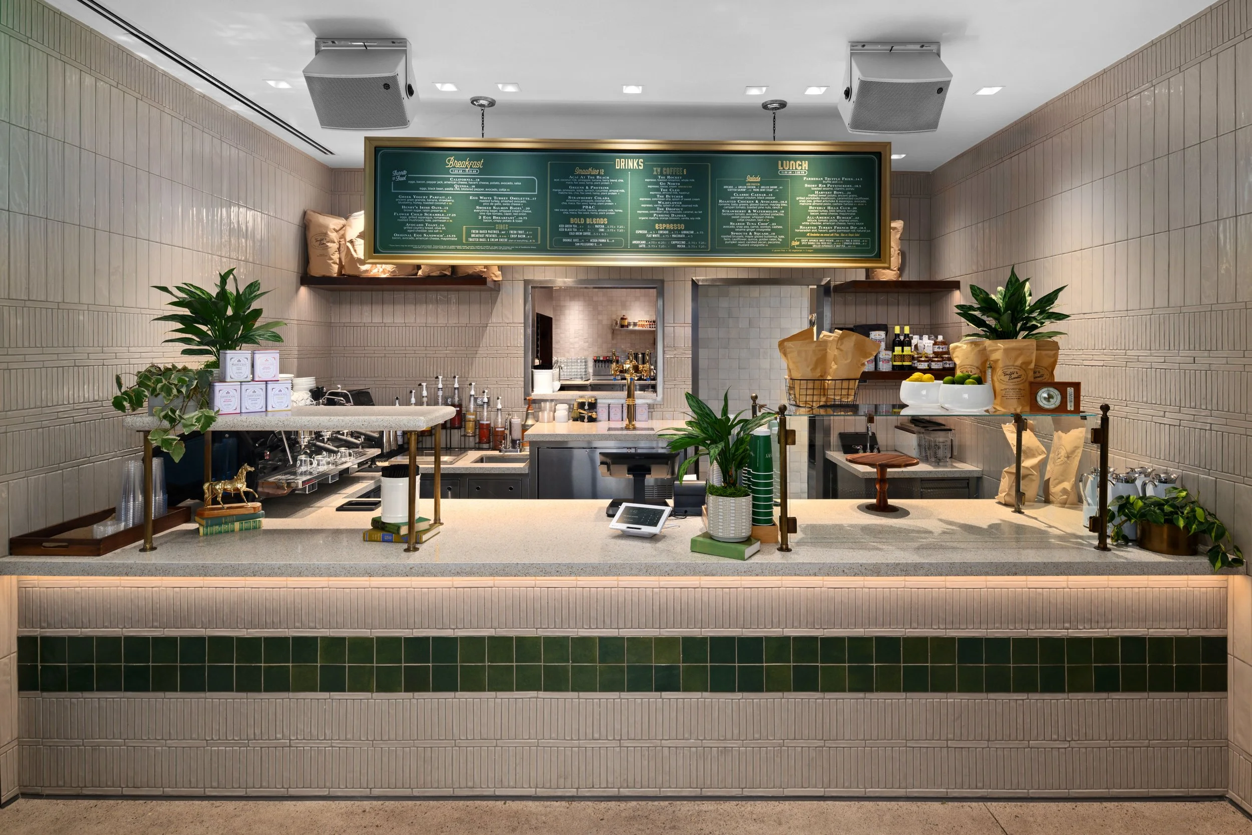 Empty cafe counter with plants, a cash register, and a menu board overhead.