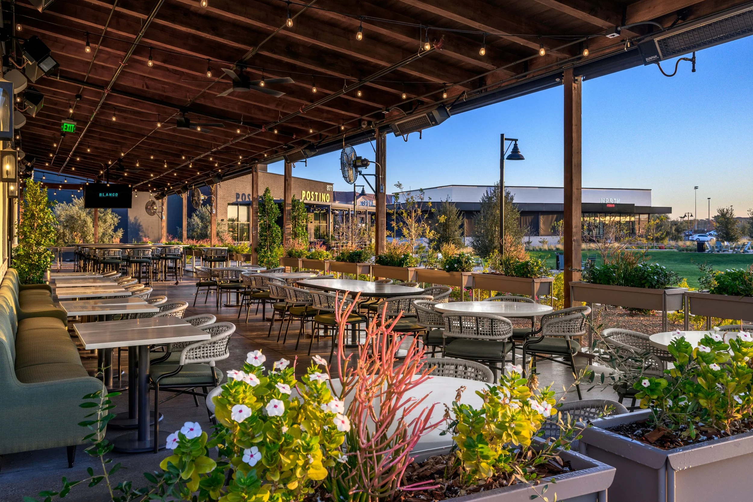 Outdoor restaurant patio with tables and chairs, surrounded by potted plants, with a view of storefronts and green lawn in the background during sunset.