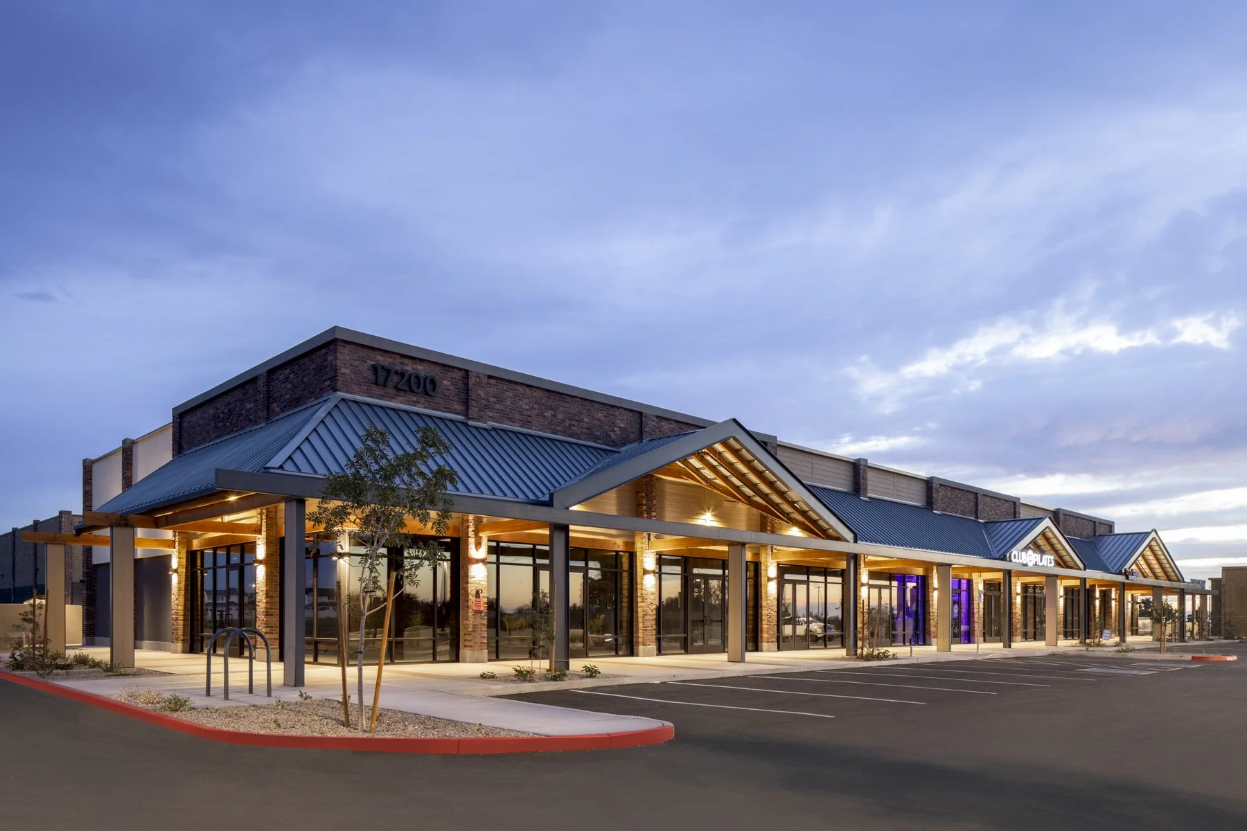 Exterior of a modern shopping center or retail store with a covered walkway and parking lot, during dusk with a cloudy sky.