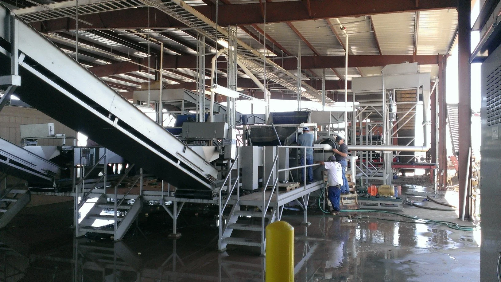 Industrial workers assembling or maintaining equipment in a large warehouse with metal stairs, pipes, and machinery.