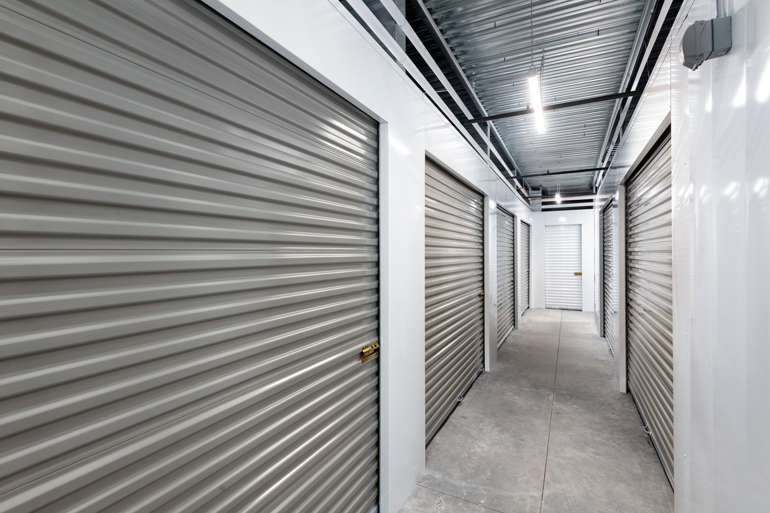 A hallway inside a storage facility with multiple metal roll-up storage unit doors and overhead lighting.