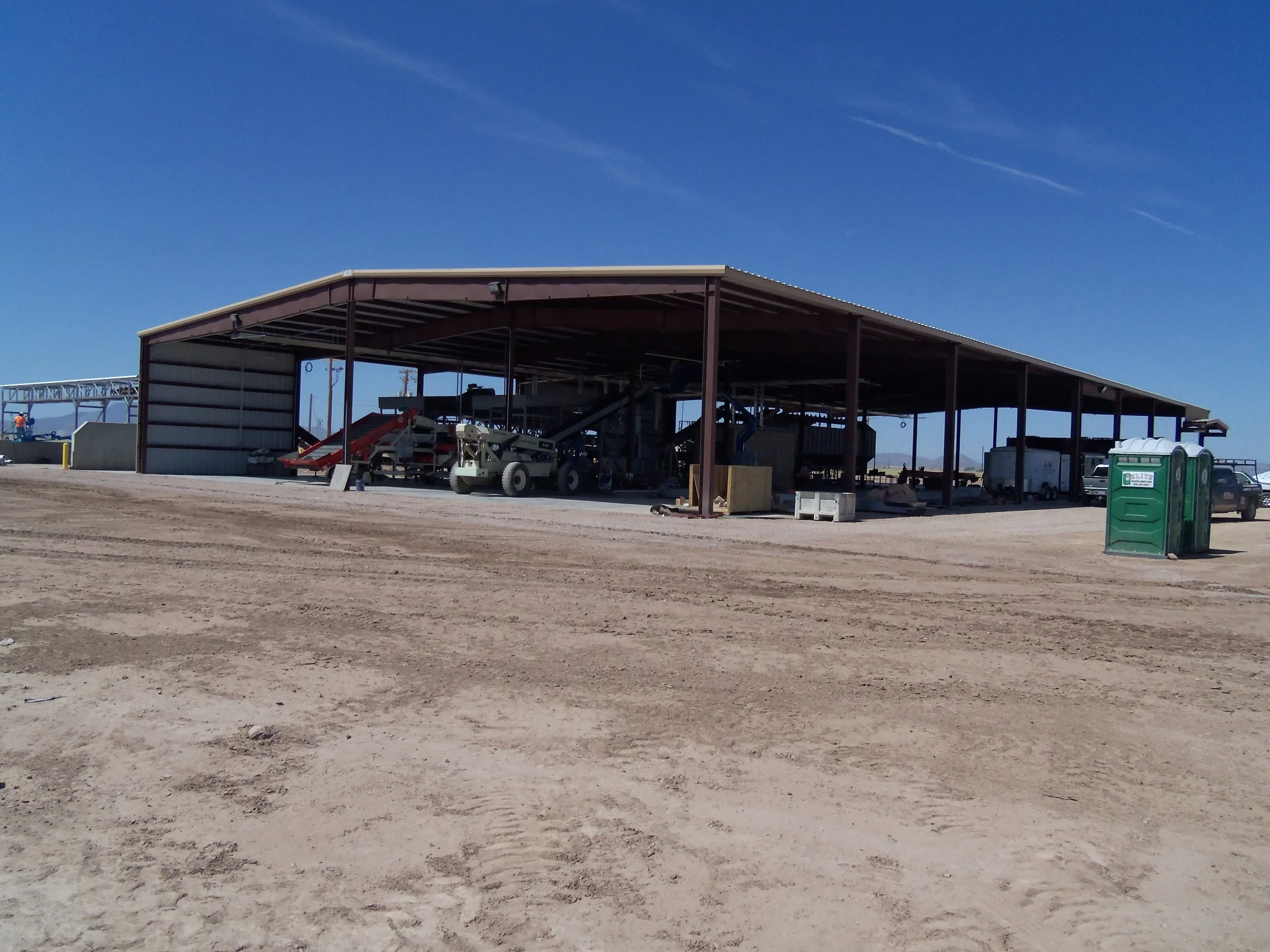 A large open-sided metal building under construction on a dirt lot, with construction equipment and trucks inside, and two portable toilets outside on the right side.