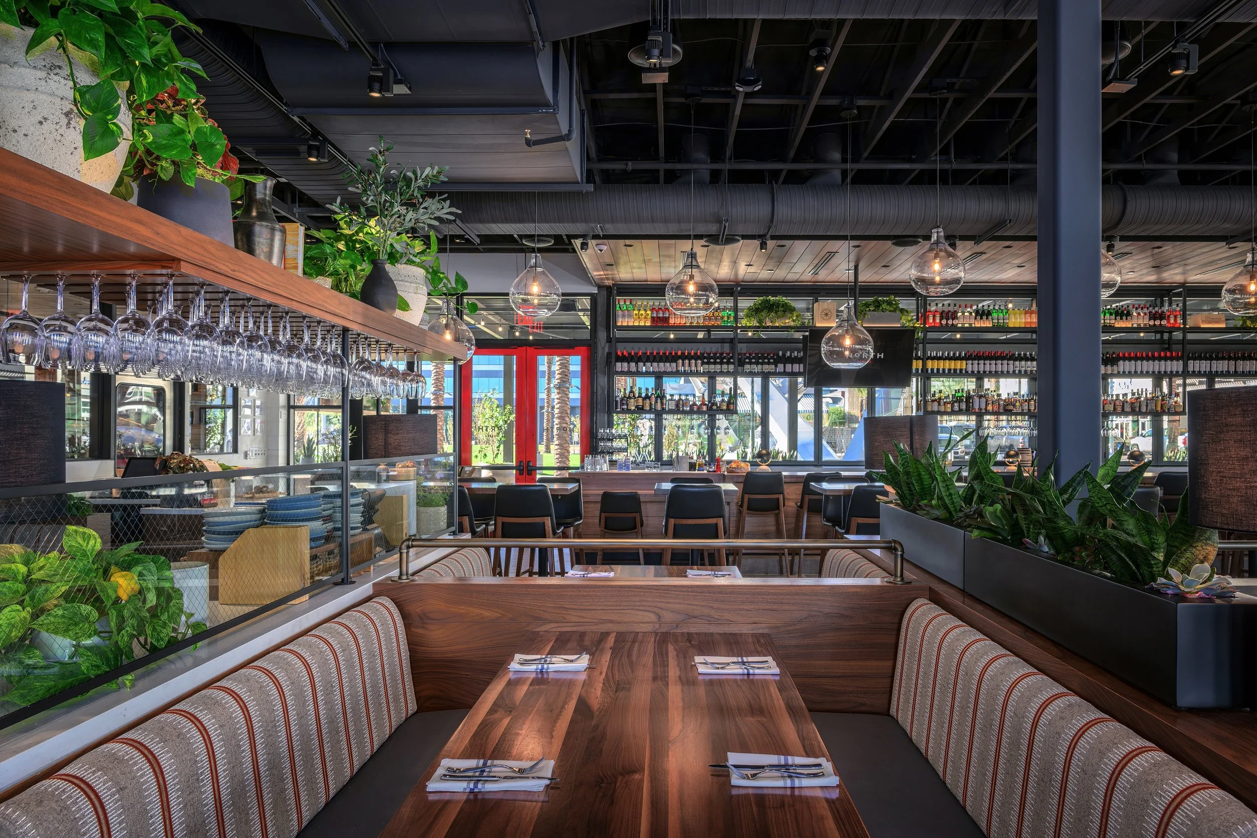 Interior of a modern restaurant with wooden tables, hanging glass light fixtures, and wine bottles on shelves in the background. The space features large windows, green plants, and a mix of seating options.