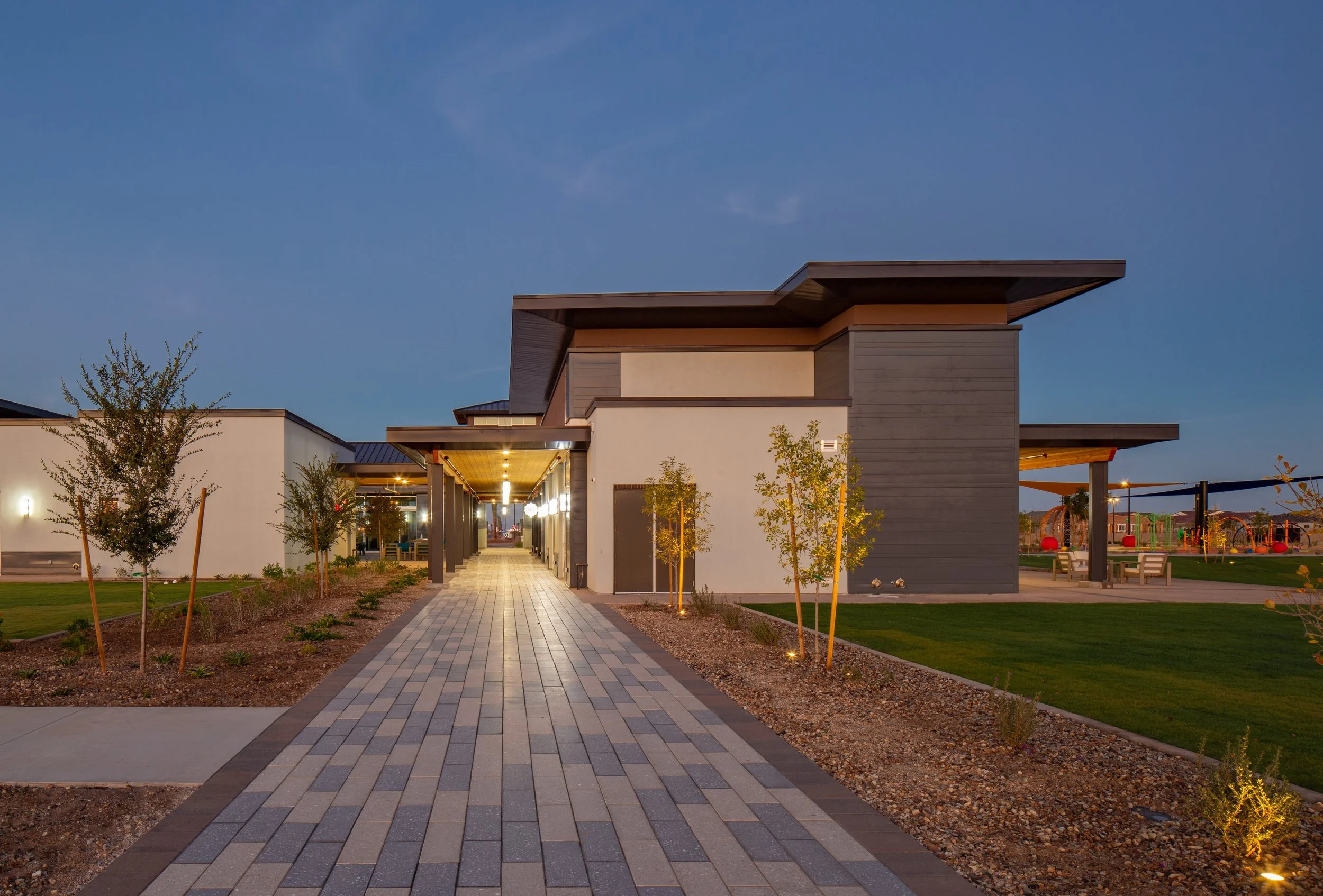 Modern building with a paved walkway, outdoor lighting, trees, and a playground in the background during evening.