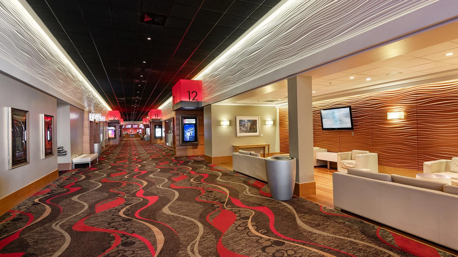 A brightly lit casino hallway with a patterned carpet featuring red, black, and beige wavy lines. There are multiple illuminated signs with numbers hanging from the ceiling, and seating areas with cream-colored sofas alongside wall-mounted screens an
