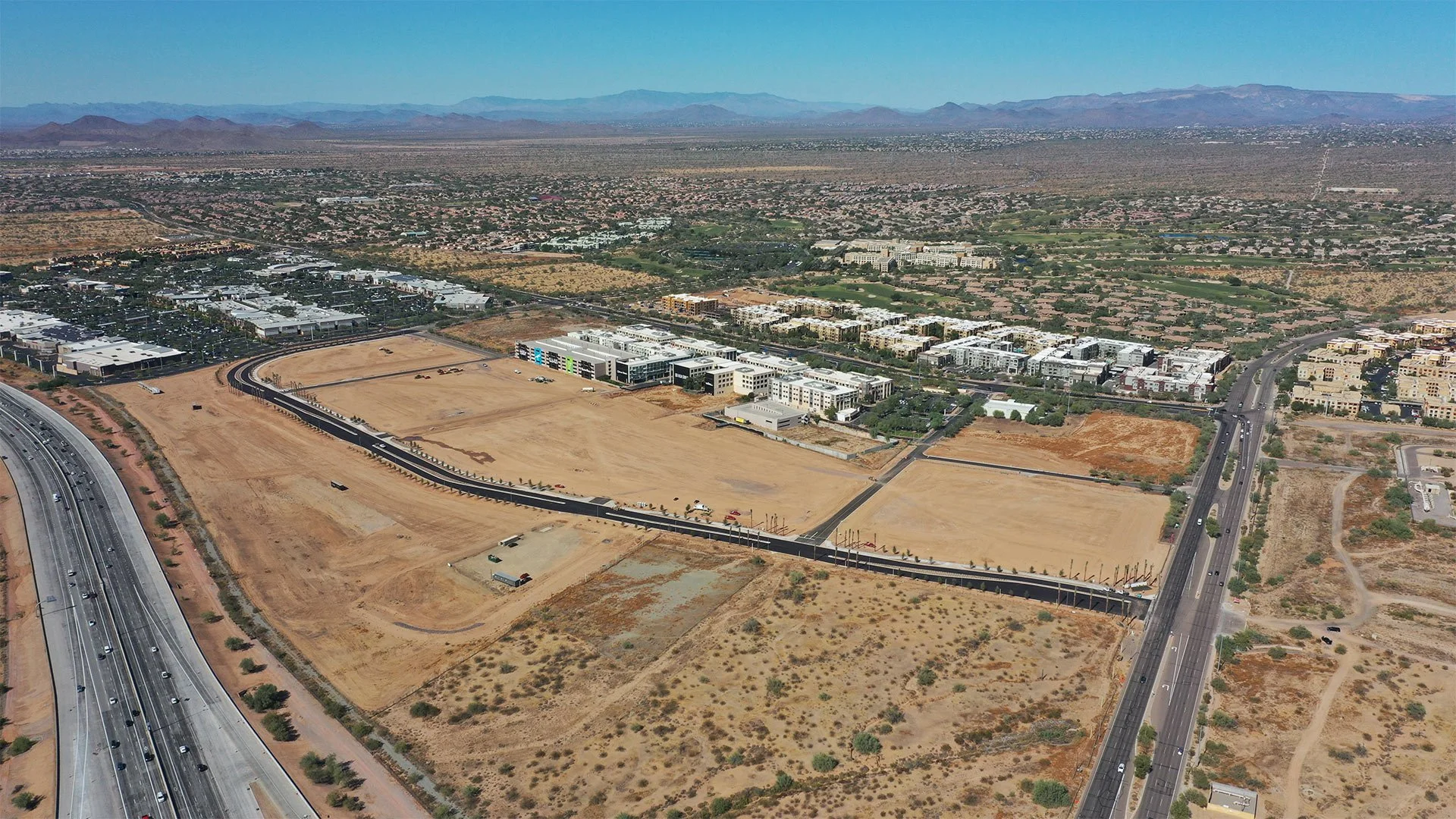 Aerial view of a construction site with cleared land, roads, and ongoing building development in a desert area near a city with mountains in the background.