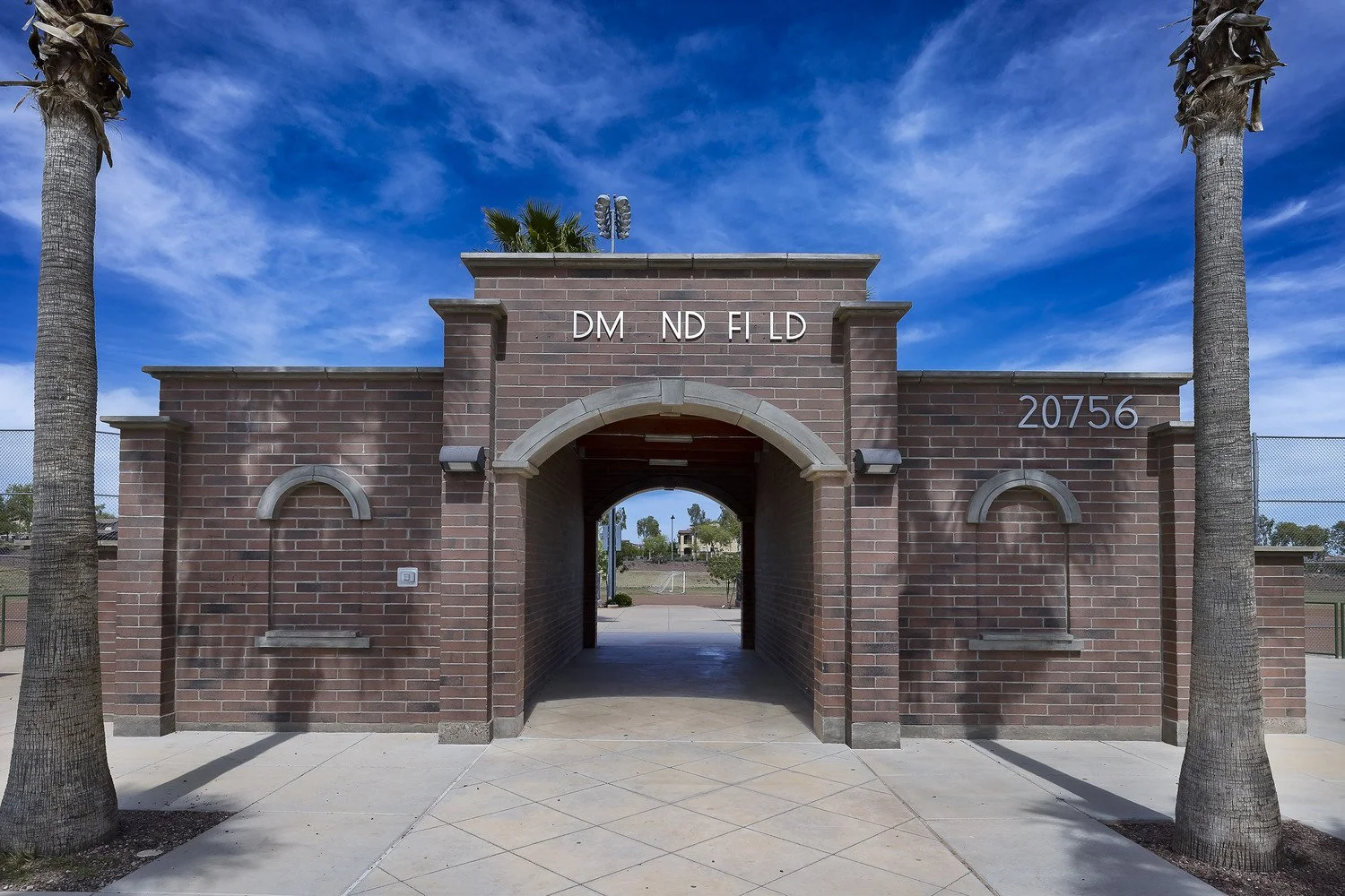 Brick structure with an arched entryway, flanked by two palm trees, leading to a school sports field, under a blue sky with wispy clouds.