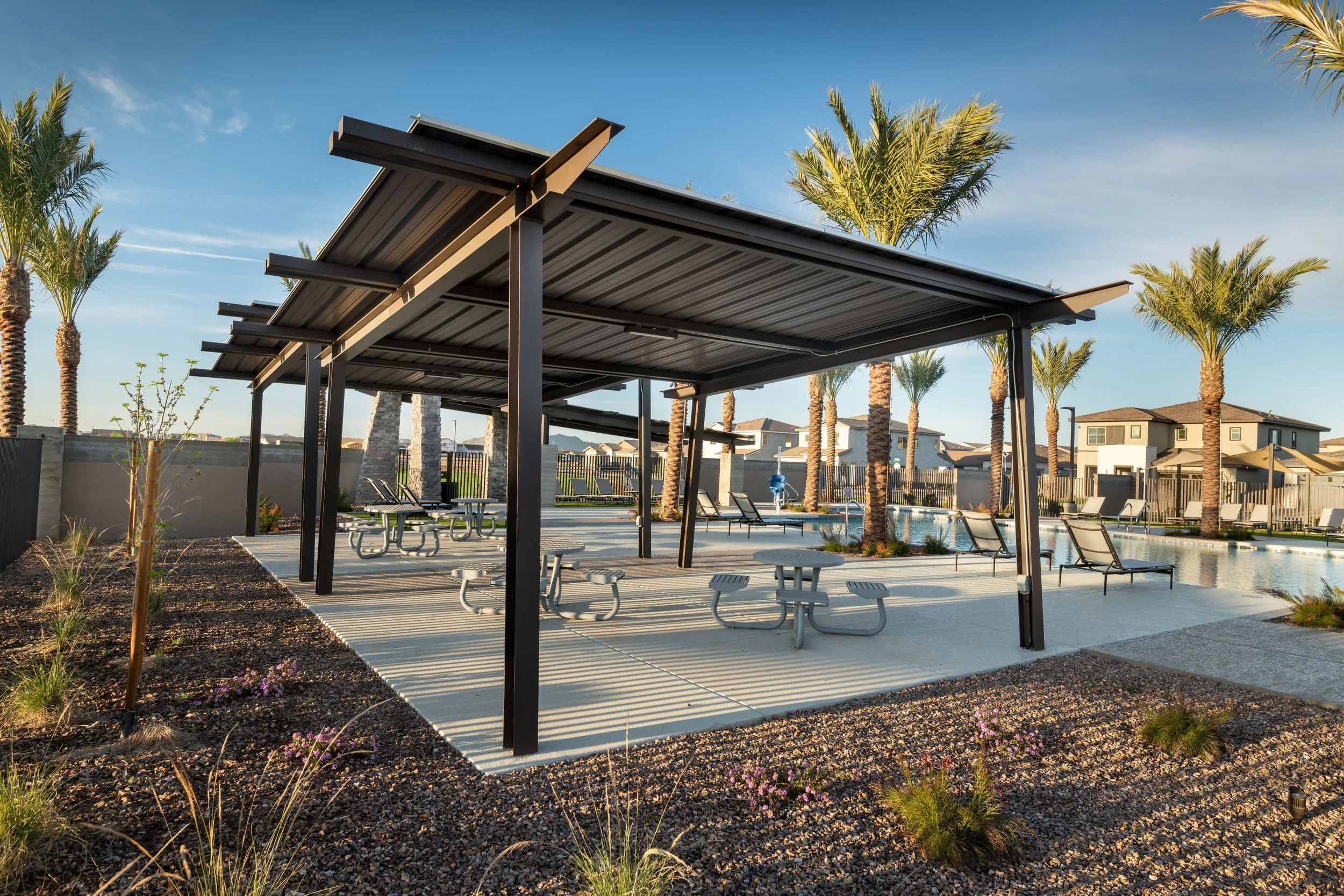 Outdoor community pool area with a covered picnic area, lounge chairs, palm trees, and a water slide in the background on a sunny day.