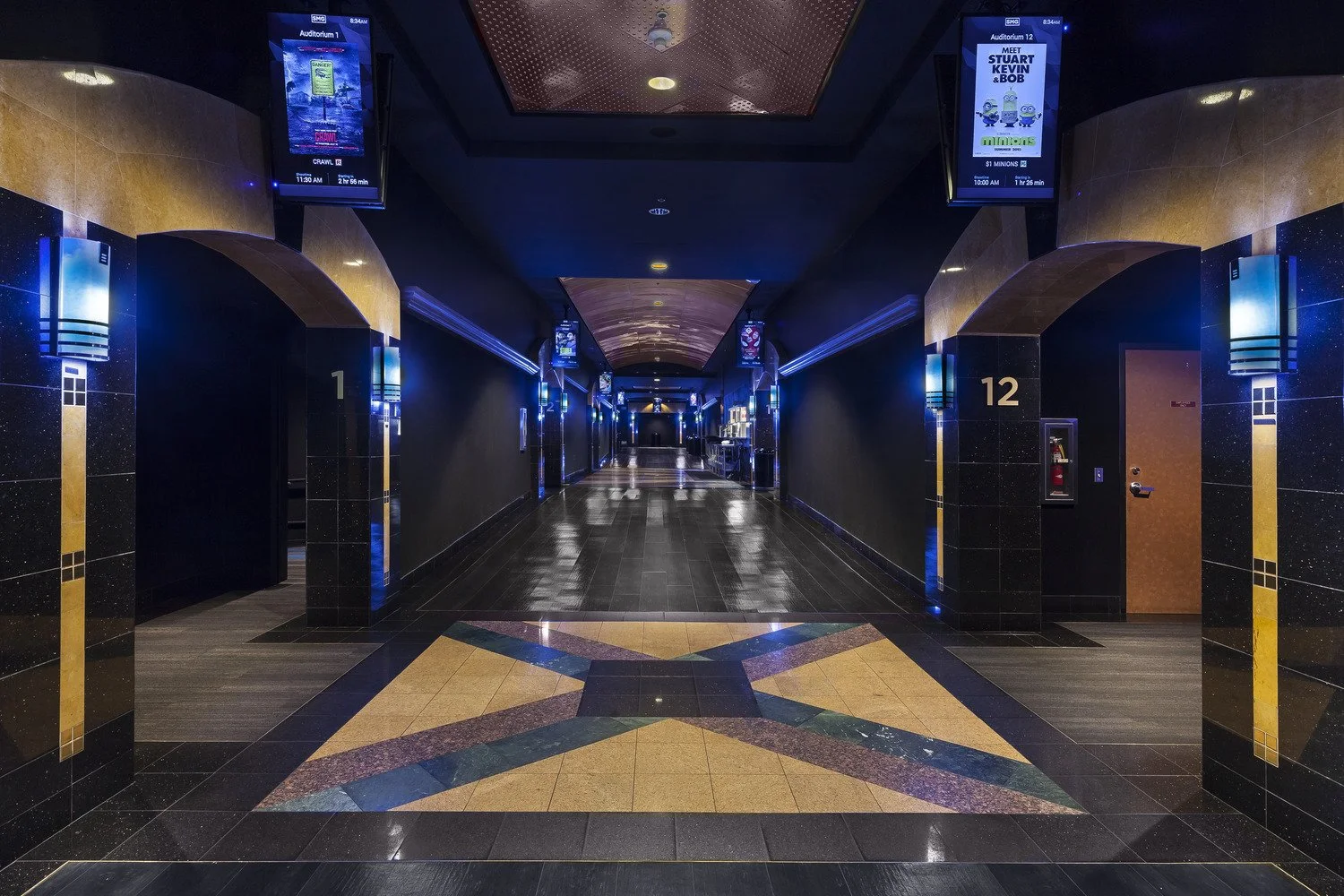 Empty movie theater hallway with arched doorways numbered 1 and 12, illuminated blue wall sconces, digital displays hanging from the ceiling, and a colorful star pattern on the tiled floor.