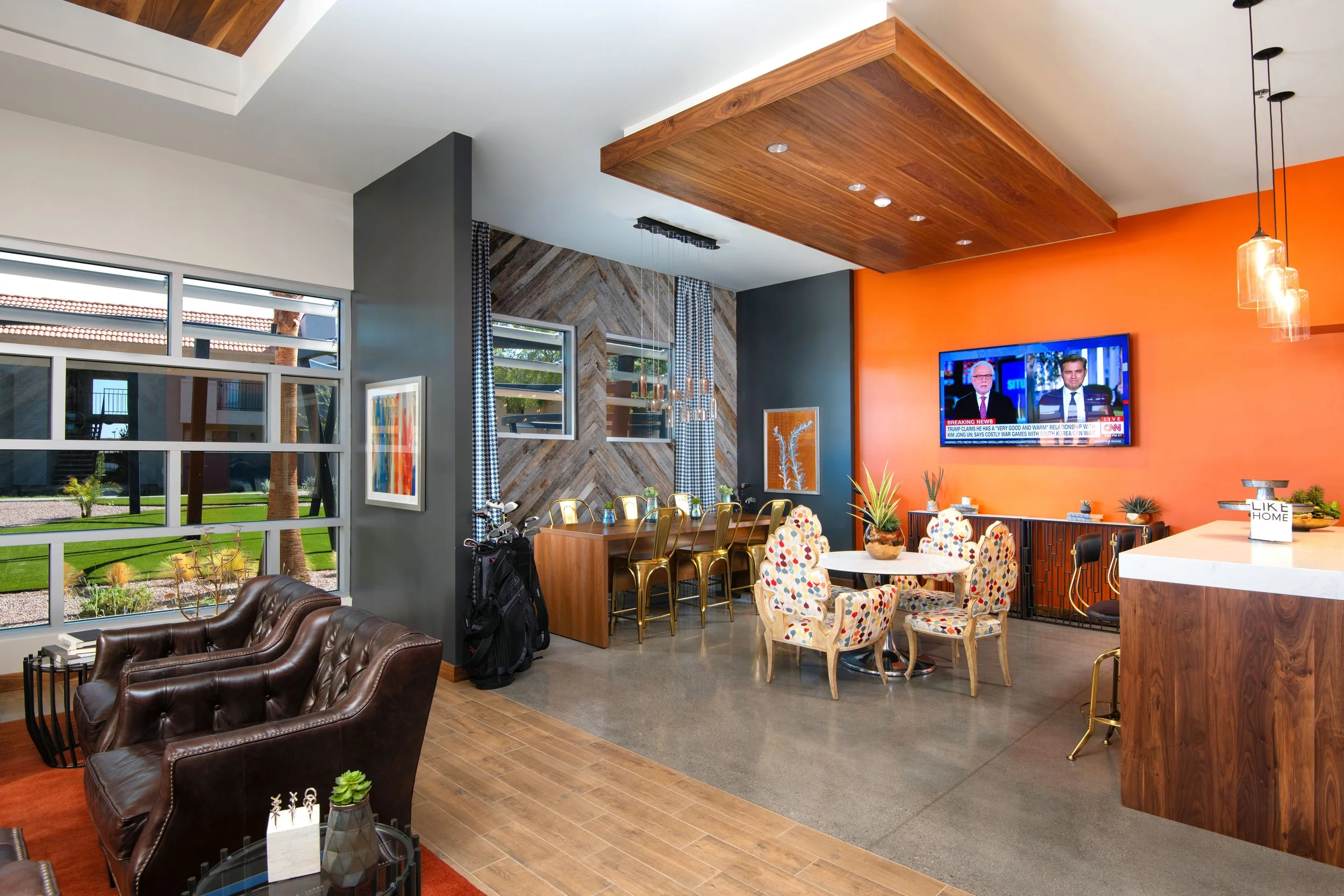 A modern living room with brown leather armchairs, a dining area with a round table and colorful patterned chairs, a television mounted on an orange wall, and large windows with a view of a green outdoor space.