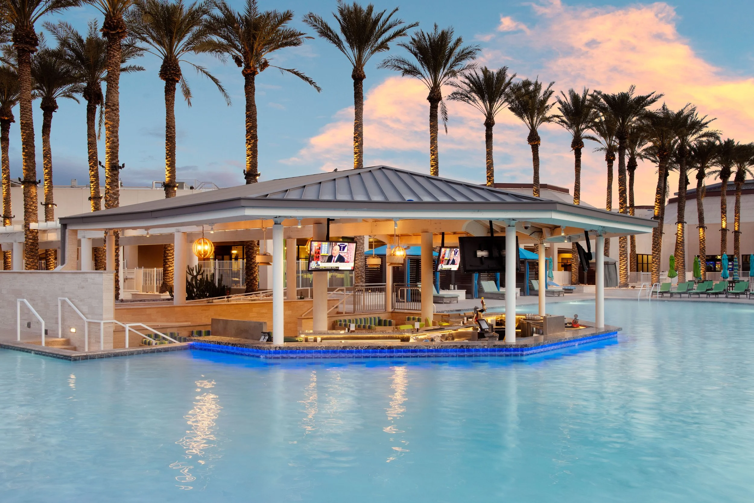 Outdoor pool area with a swim-up bar, palm trees, lounge chairs, and a sunset sky in the background.