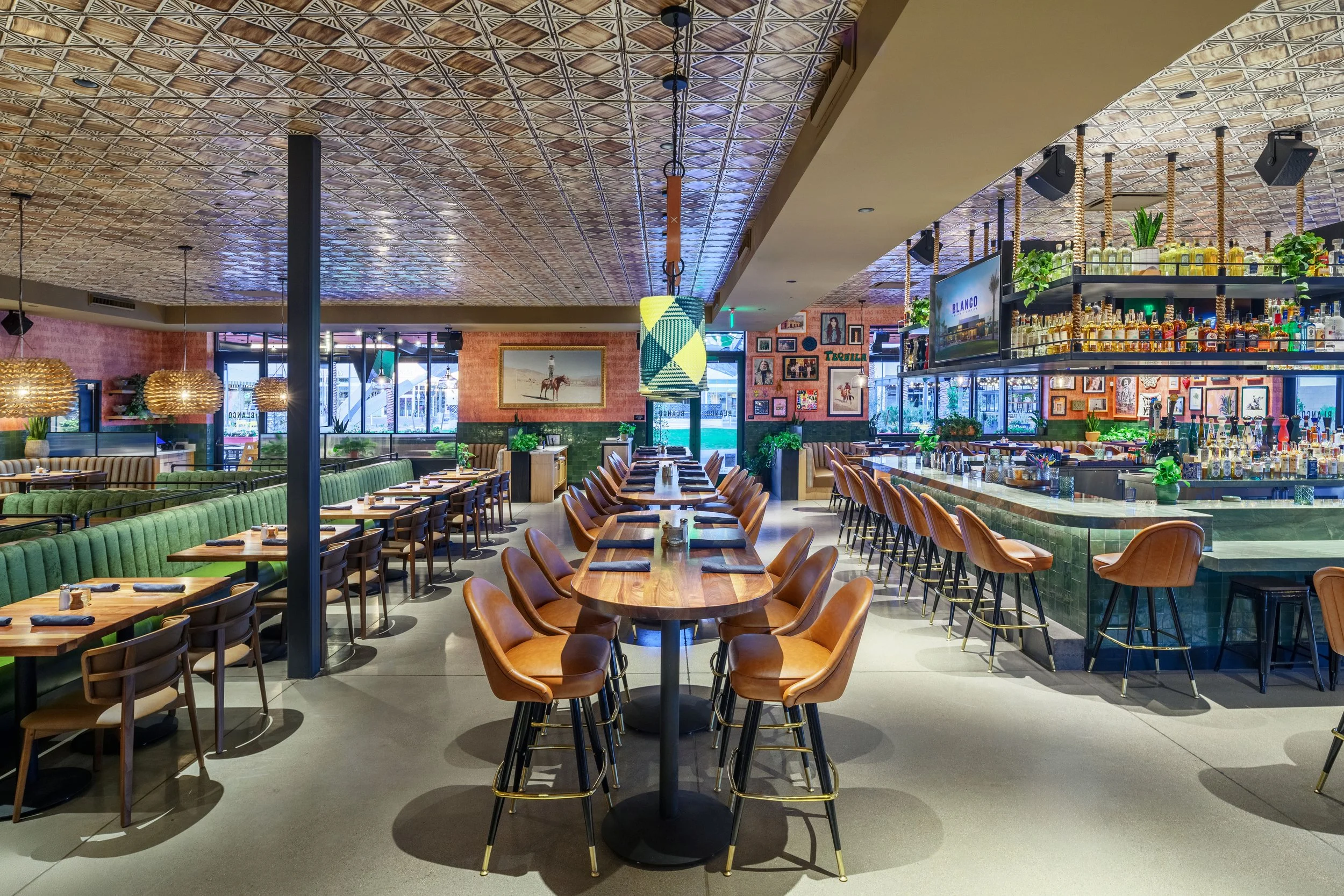 Interior of a modern restaurant with a bar on the right, featuring leather bar stools and shelves of drinks. In the center, a large communal wooden table with leather chairs. To the left, seating with green upholstered banquettes and small wooden tab