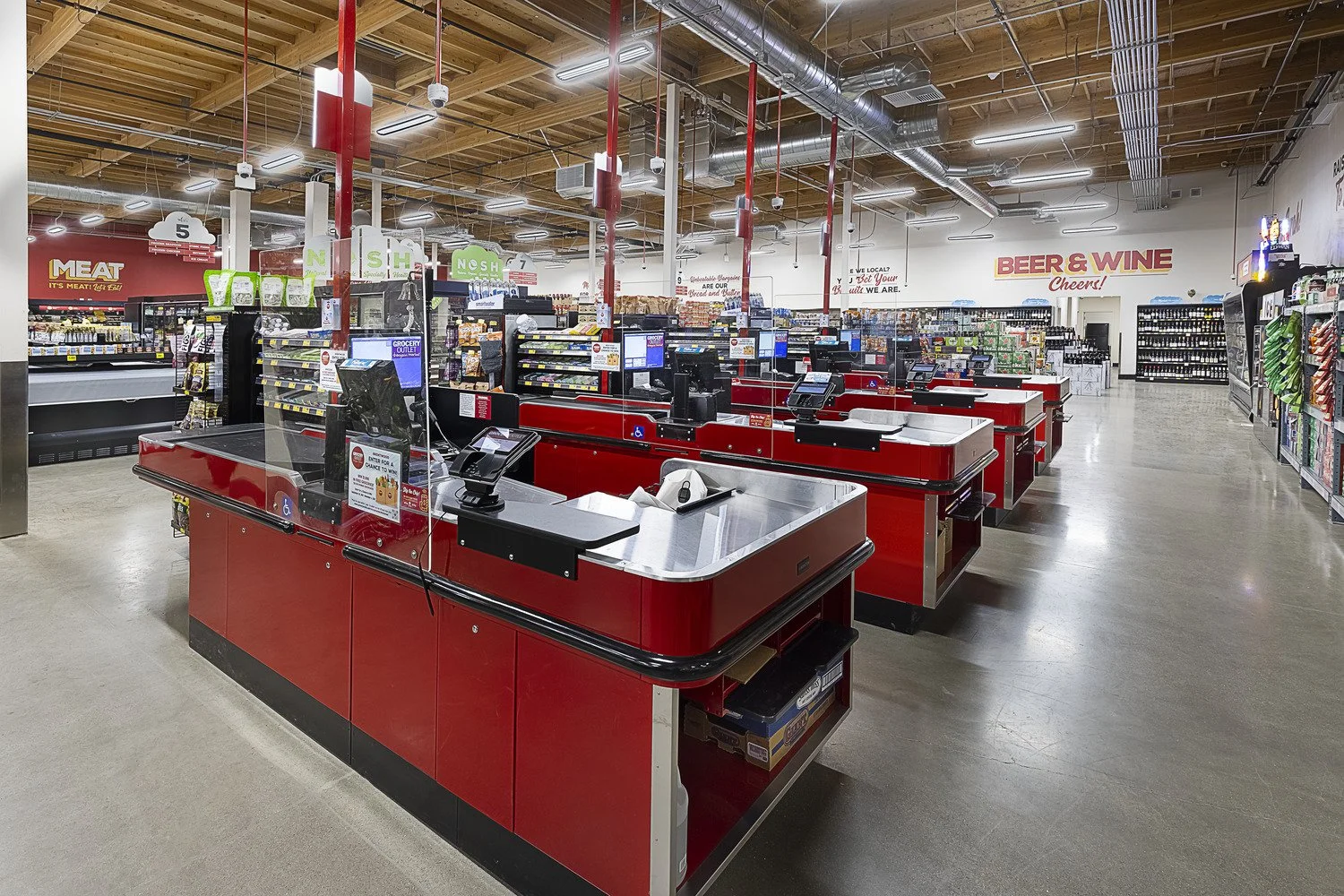 Empty checkout counters with red counters, point-of-sale screens, and checkout lanes in a grocery store with aisle signs for meat, produce, and beer & wine.