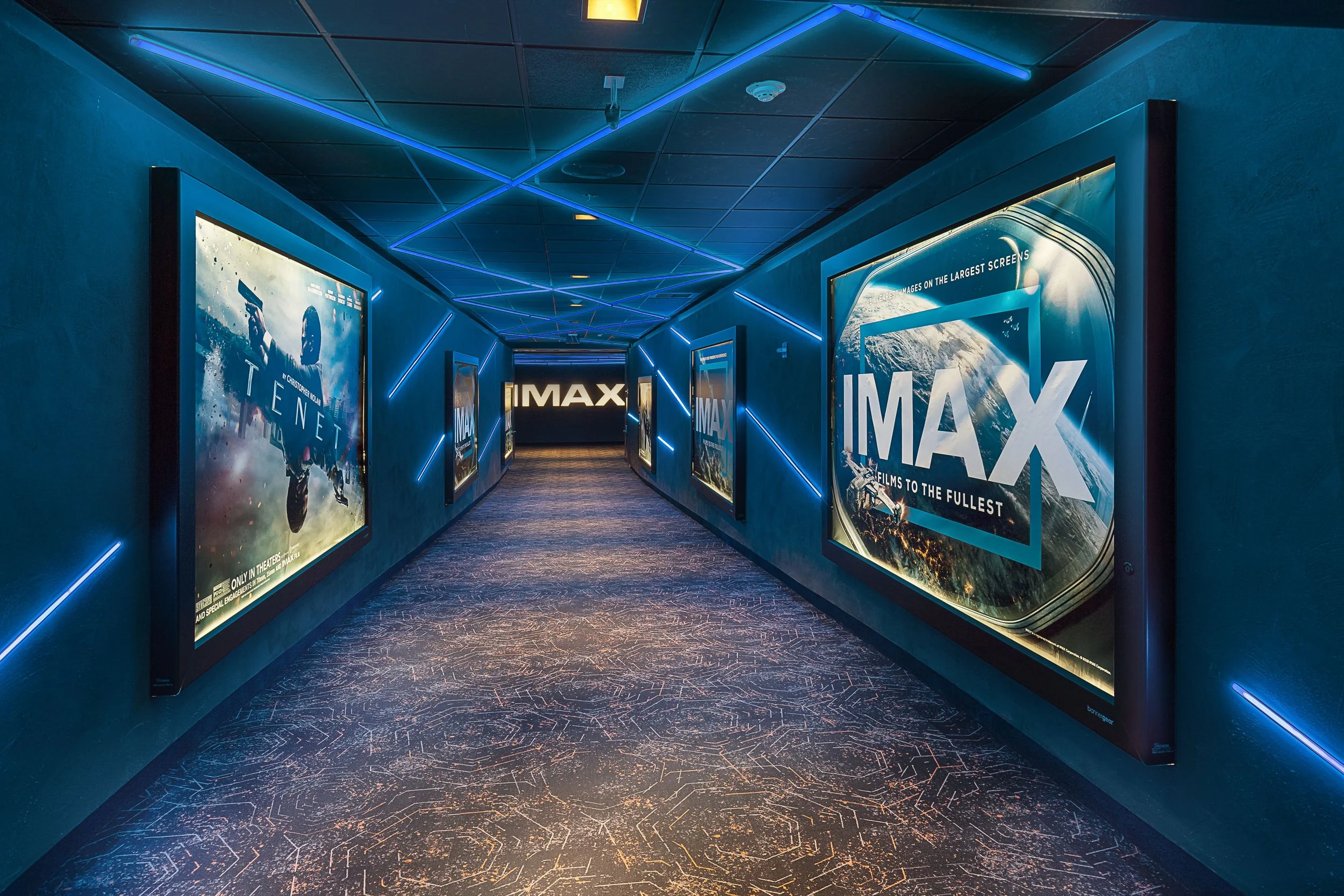 Movie theater hallway with blue walls, illuminated by blue neon lights on the ceiling and walls, displaying posters for IMAX films, leading to an IMAX screen at the end of the corridor.