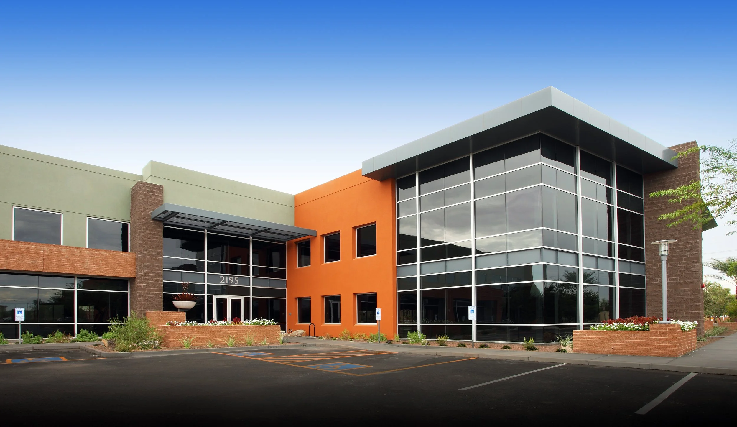 Modern commercial building with large glass windows, orange and green exterior walls, landscaped plants, and parking lot in front.