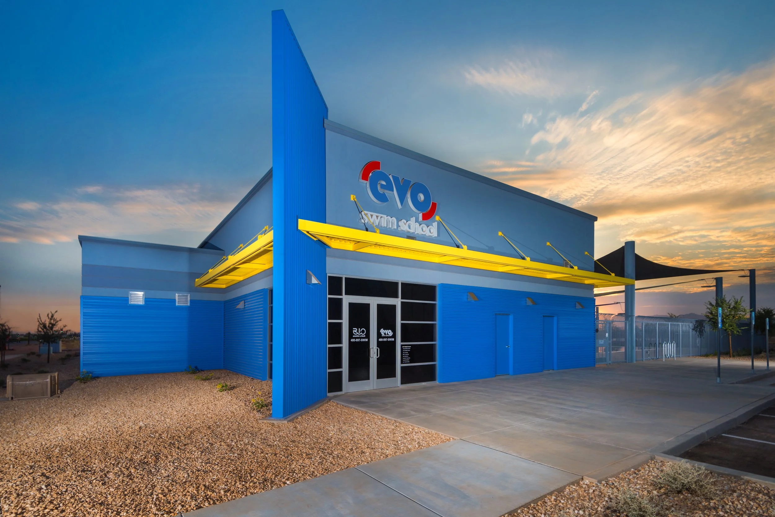 Blue building with yellow awnings at sunset, with signs for Evo Swim School and a fenced outdoor area.
