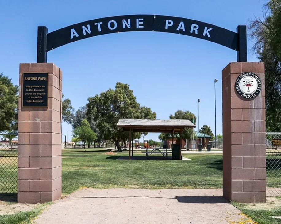 Entrance to Antone Park with brick pillars and a black arch sign, a picnic table under a gazebo, and trees in the background.