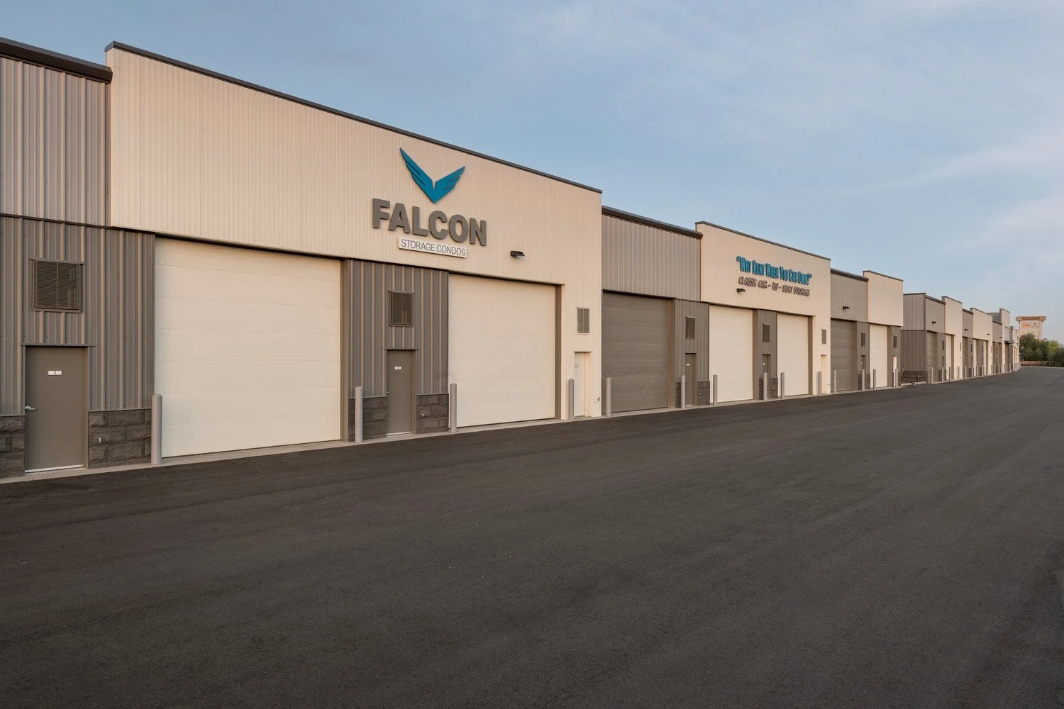 A row of industrial storage units with roll-up doors on a paved lot. The warehouse building has signage for FALCON Storage Condos and additional business names. The sky is clear.