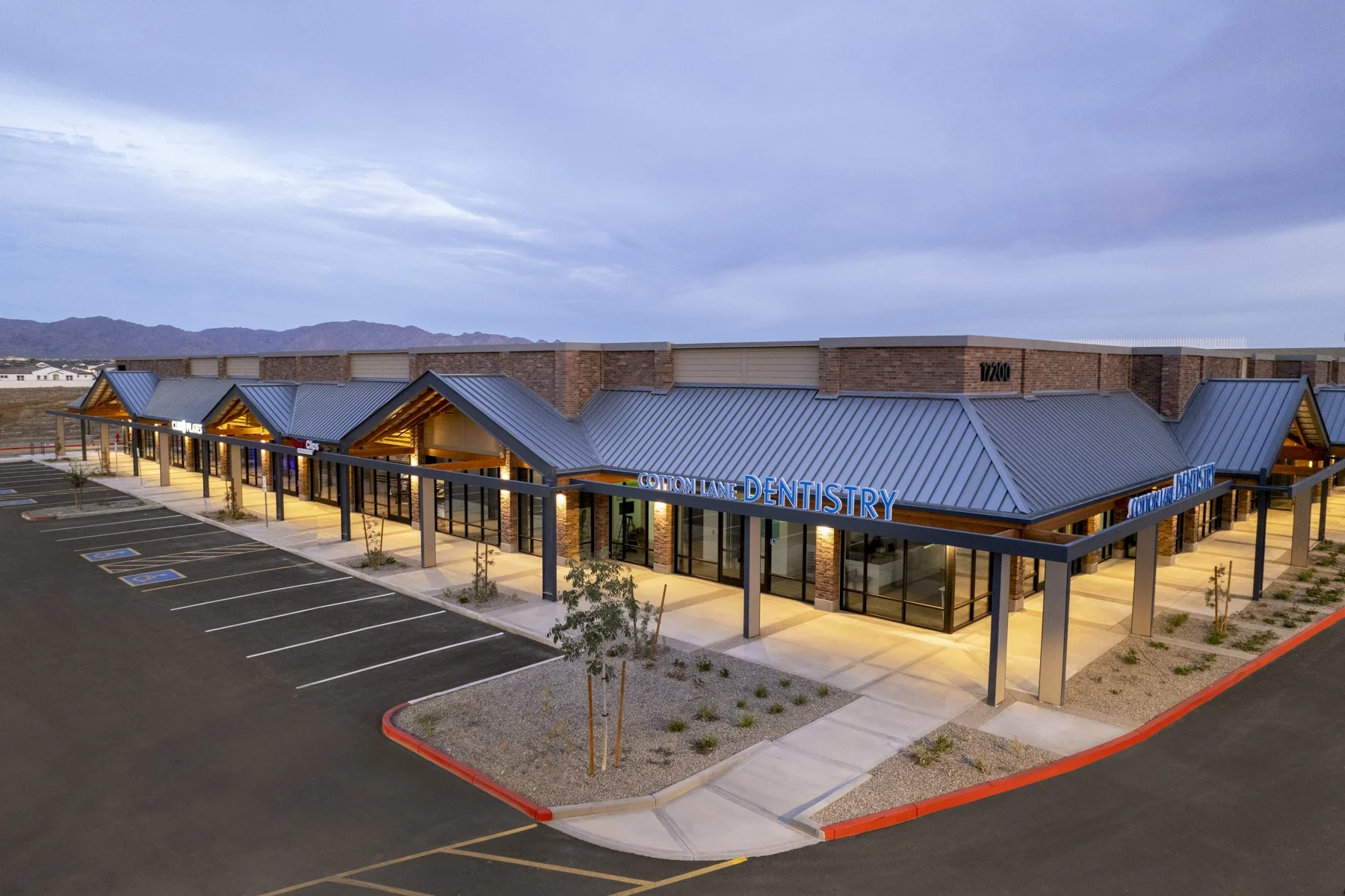 A modern shopping plaza with a dental office named Cotton Lane Dentistry, featuring a brick and metal roof design, parking lot with handicap spaces, and a sidewalk with small trees, under a cloudy sky.