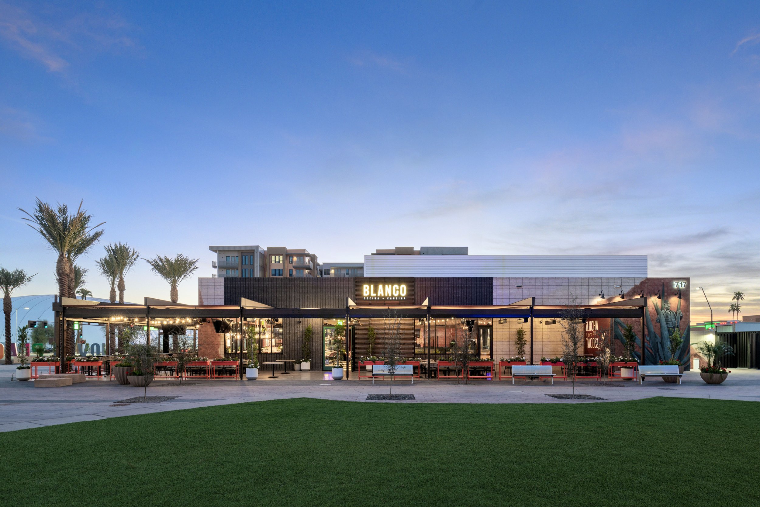 Front view of a modern restaurant named Blango with outdoor seating, surrounded by palm trees, at dusk, with a grassy area in the foreground.