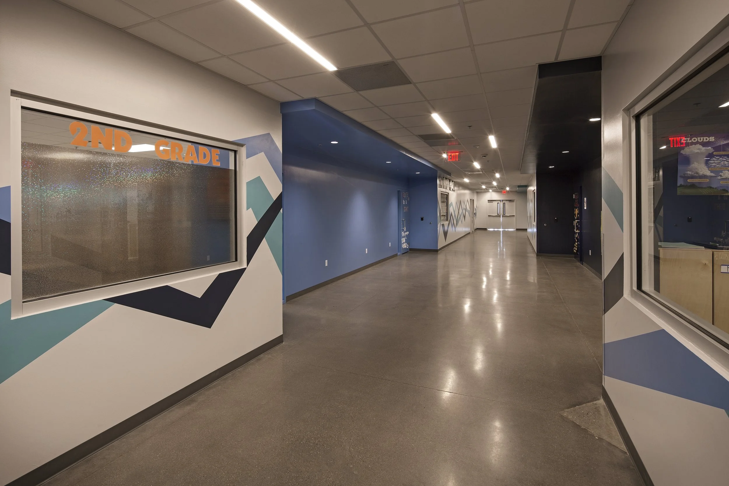Empty school hallway with blue walls, geometric wall art, and glass window on the left marked '2ND GRADE'.