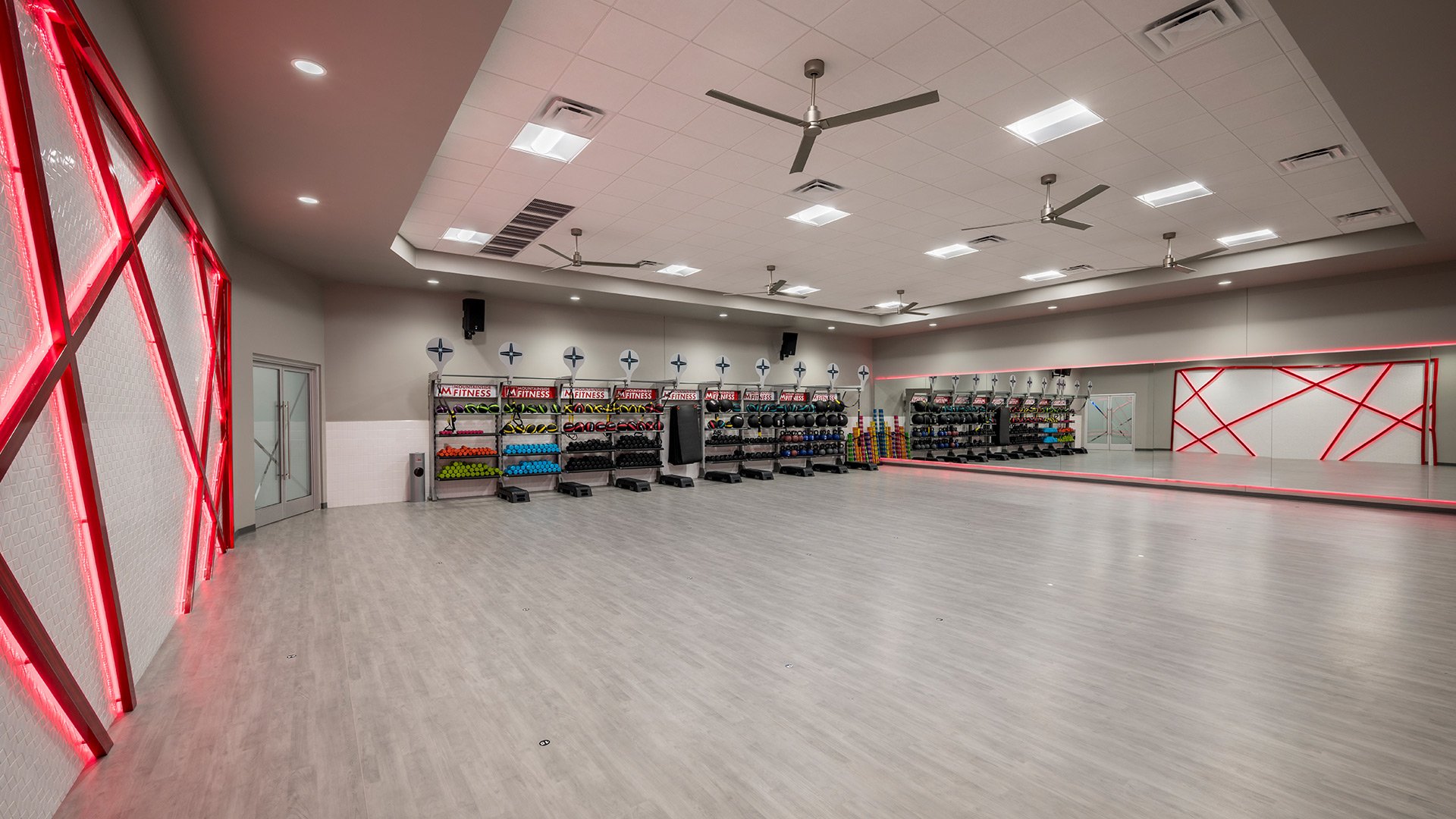 Empty fitness studio featuring wall mirrors, shelves with fitness equipment like dumbbells and medicine balls, a large mirror with red decorative borders, and ceiling fans.