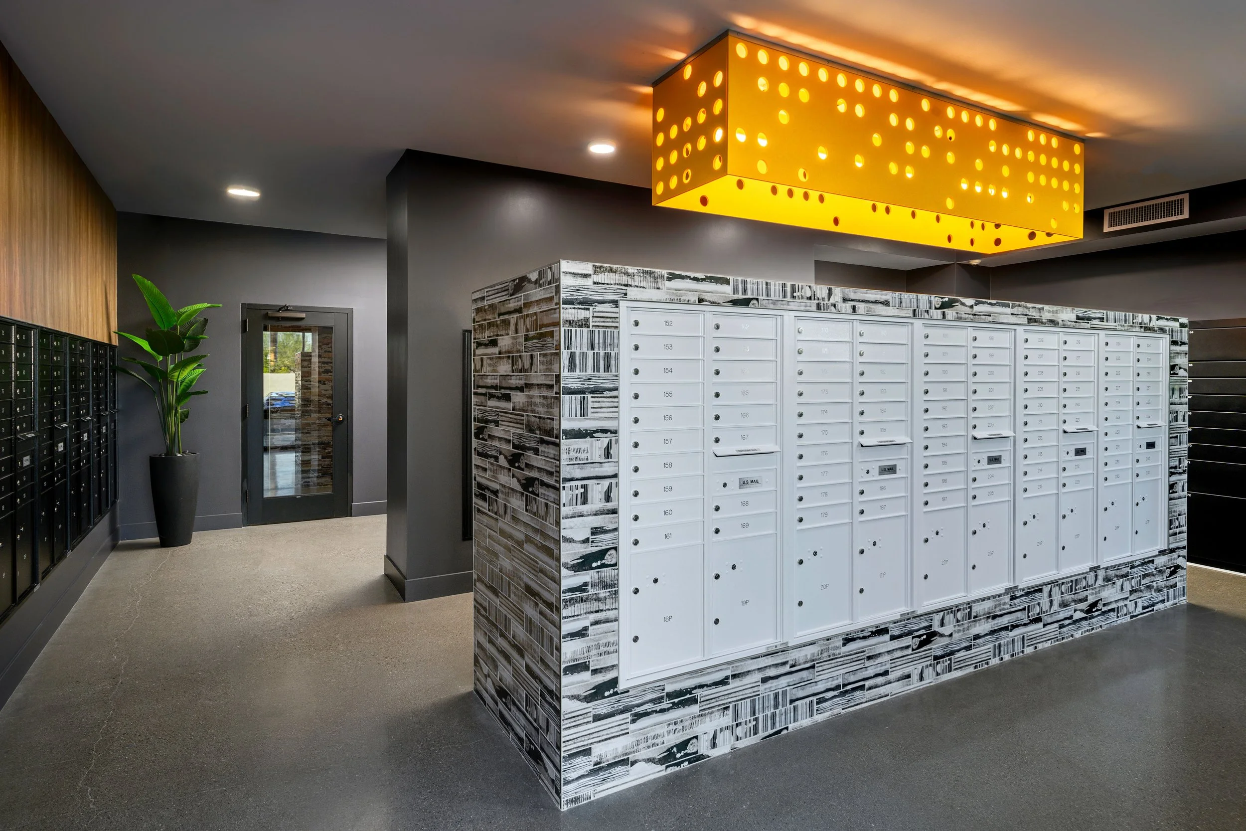 Apartment mailbox area with multiple white mailboxes on a decorative wall, a yellow ceiling light fixture, black mailboxes on the wall, a door with a window, and a potted plant.