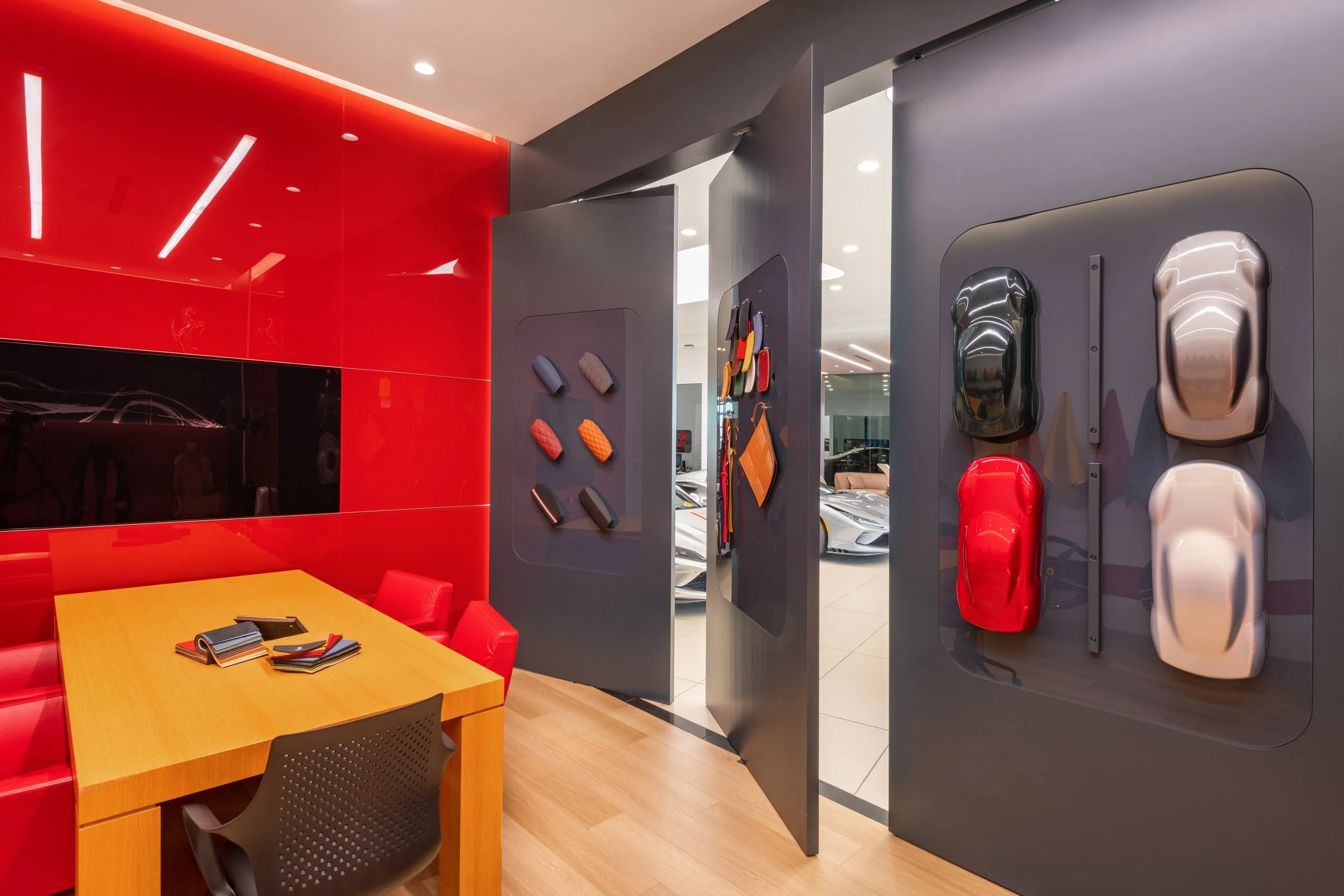 A modern showroom with a red wall and a wooden table surrounded by red and black chairs. The table has fabric samples on it.