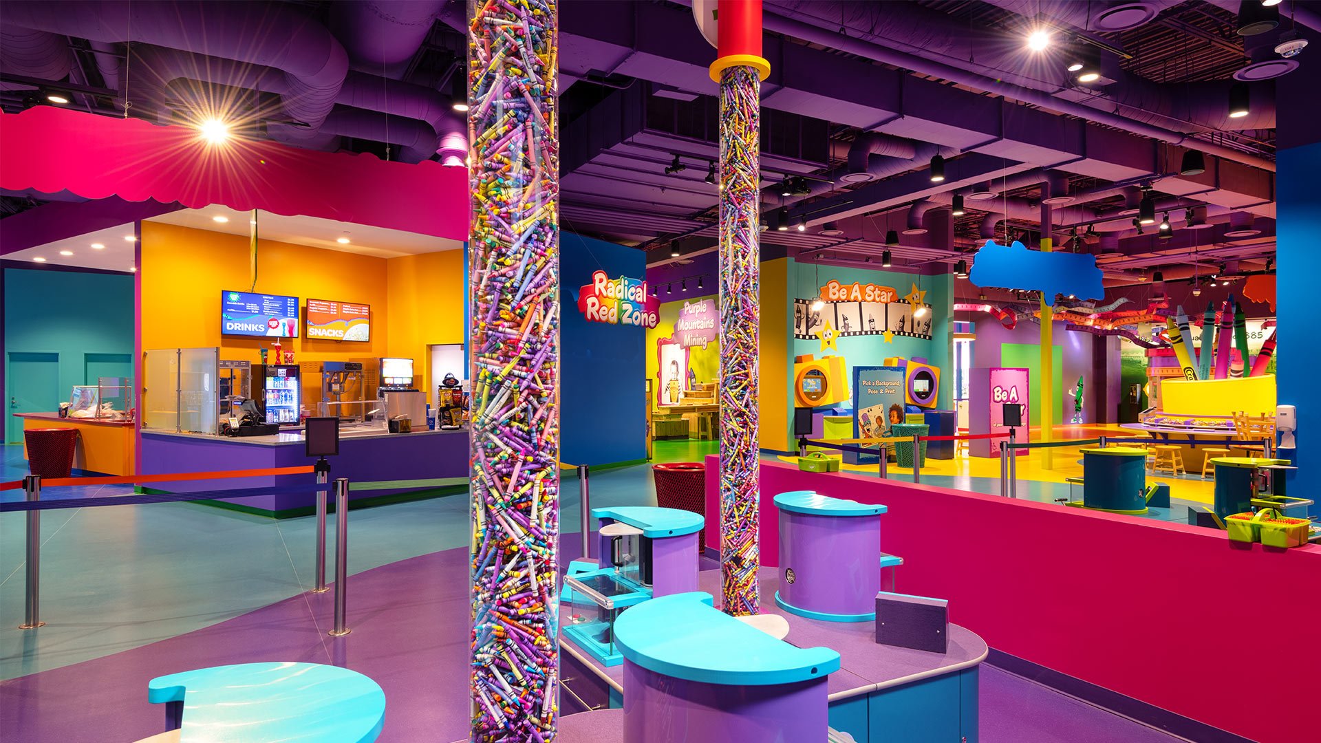 Colorful indoor play area with a snack counter, interactive stations, and vibrant decor including columns filled with plastic toys and a ceiling with pipes and lighting.