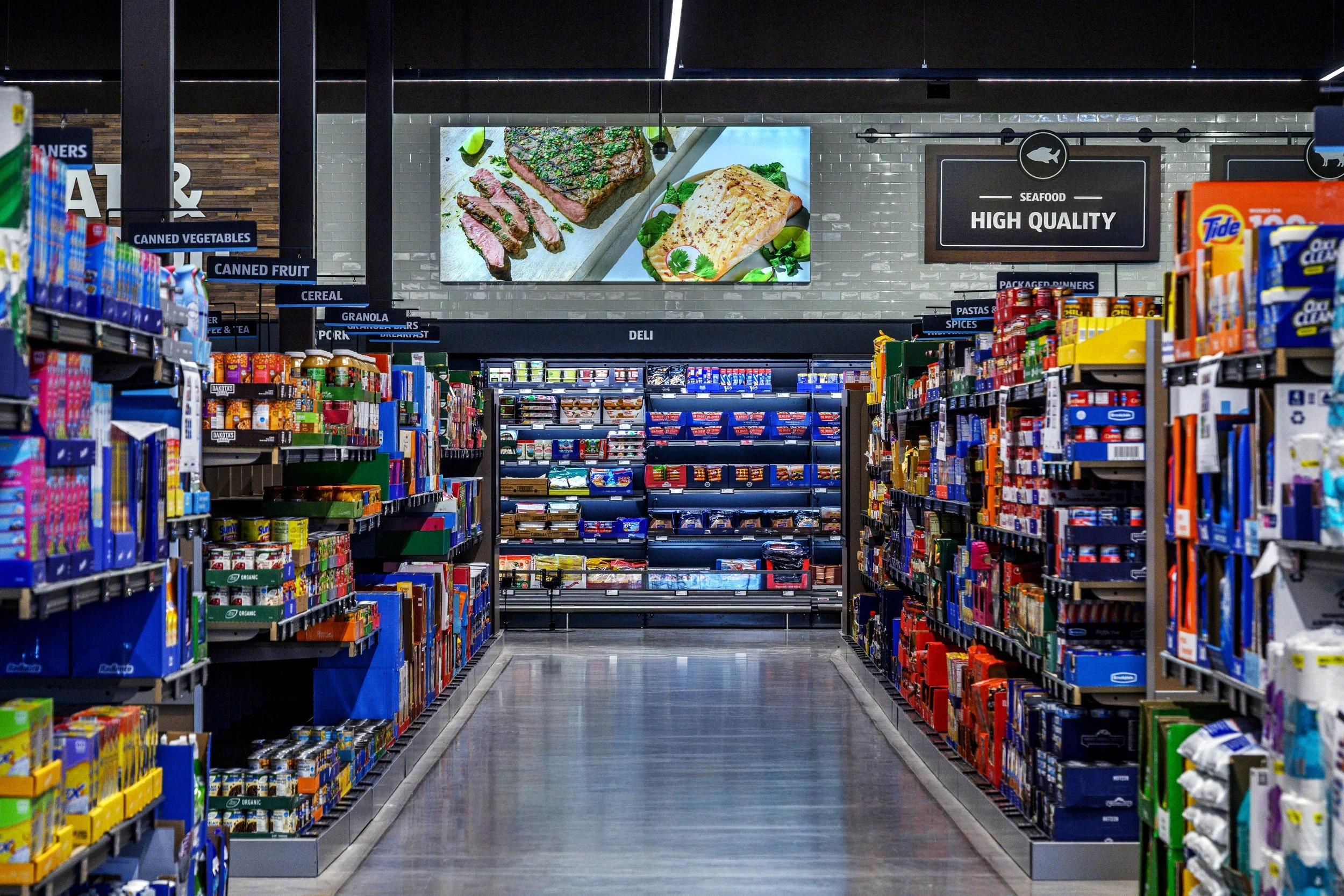 Aisle in a grocery store with shelves stocked with canned vegetables, canned fruits, cereal, pasta and spices, and a digital screen displaying images of steak and fish above. Signage indicates sections such as 'Canned Vegetables,' 'Canned Fruit,' and