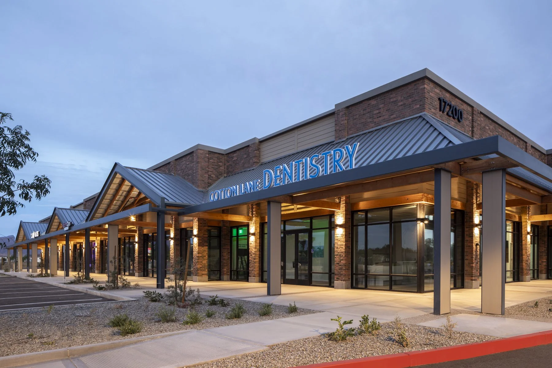 Exterior of a modern brick building with a sign that reads Cotton Lane Dentistry, with a parking lot in front and small landscaped plants.