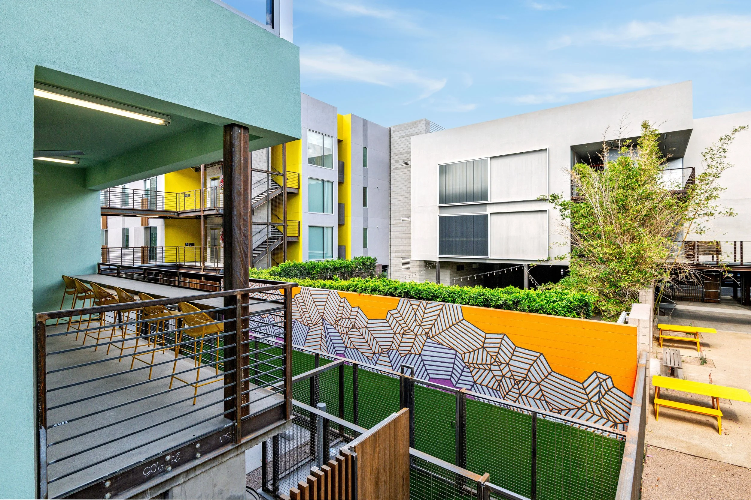 Colorful modern apartment complex with outdoor terrace, painted wall with geometric mural, trees, and benches.